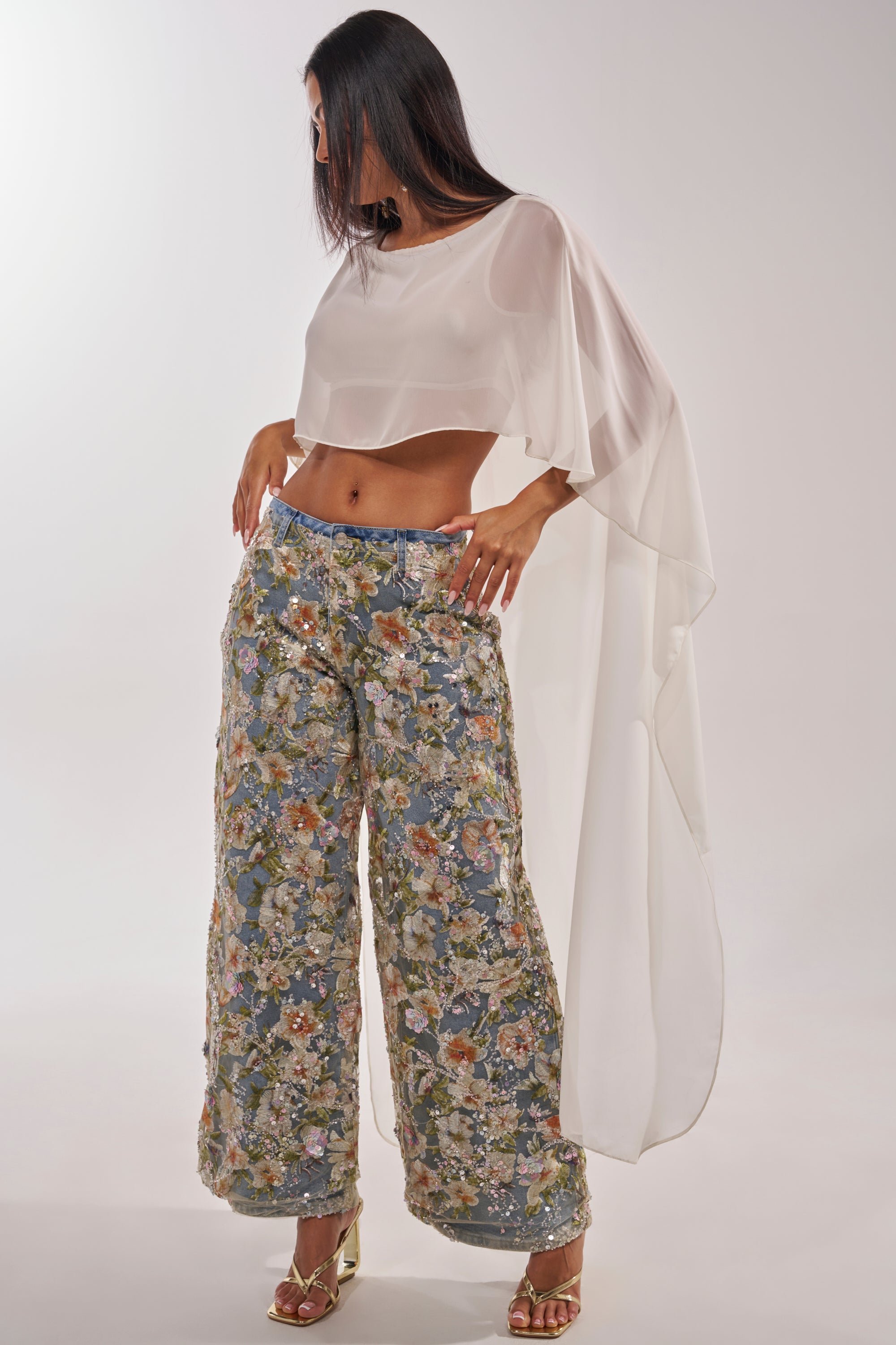A woman poses against a plain light background in IRIS RELAXED DENIM WITH FLORAL OVERLAY, paired with a sheer white asymmetrical crop top and gold heels. 4