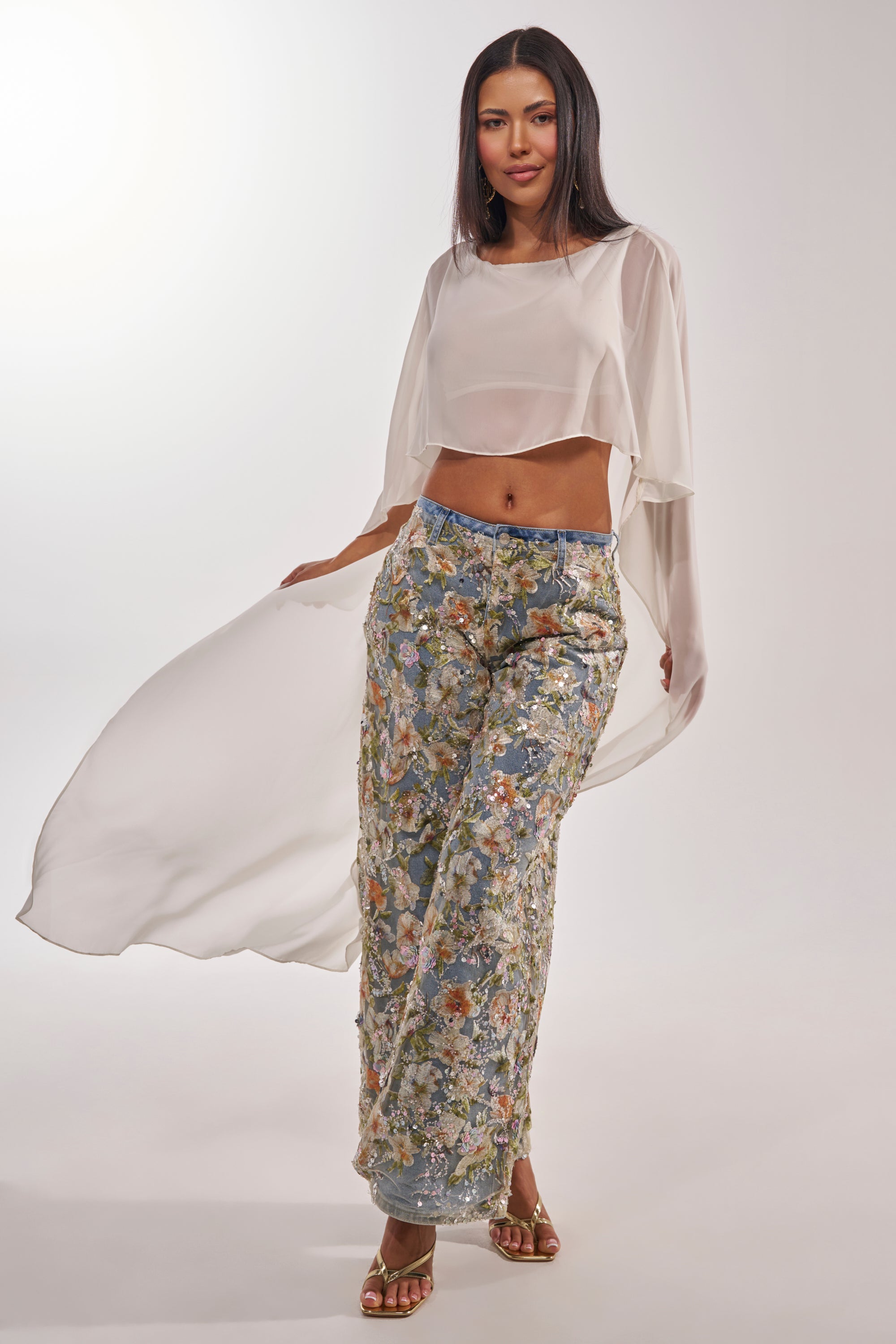 A woman poses confidently in IRIS RELAXED DENIM WITH FLORAL OVERLAY and a sheer white long-sleeve crop top, complemented by gold sandals and flowing fabric details. She has long dark hair and stands against a plain white background. 5