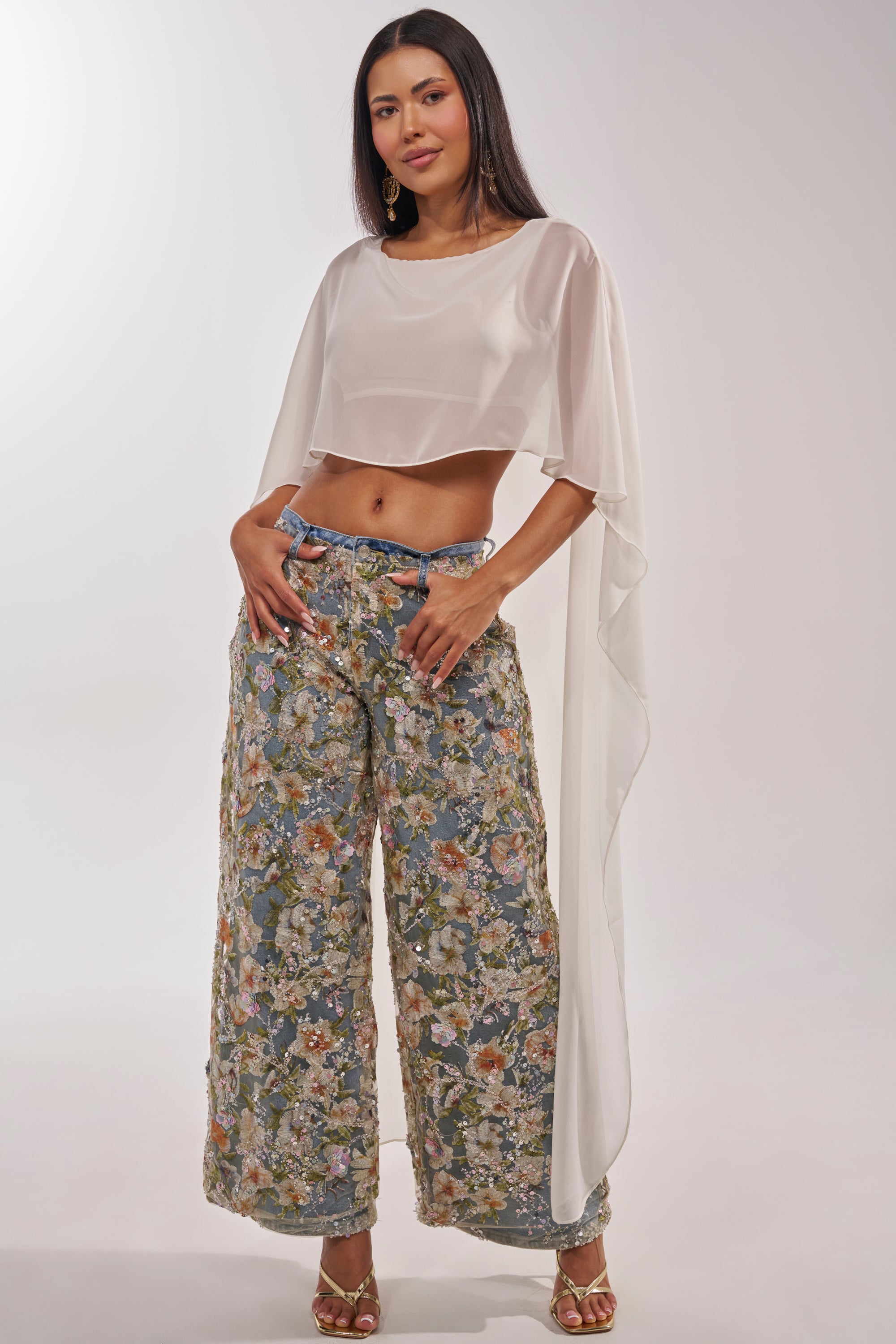 A woman poses confidently in IRIS RELAXED DENIM WITH FLORAL OVERLAY, paired with a sheer white cropped top and gold heels. She has long straight hair and stands against a plain light background. 6
