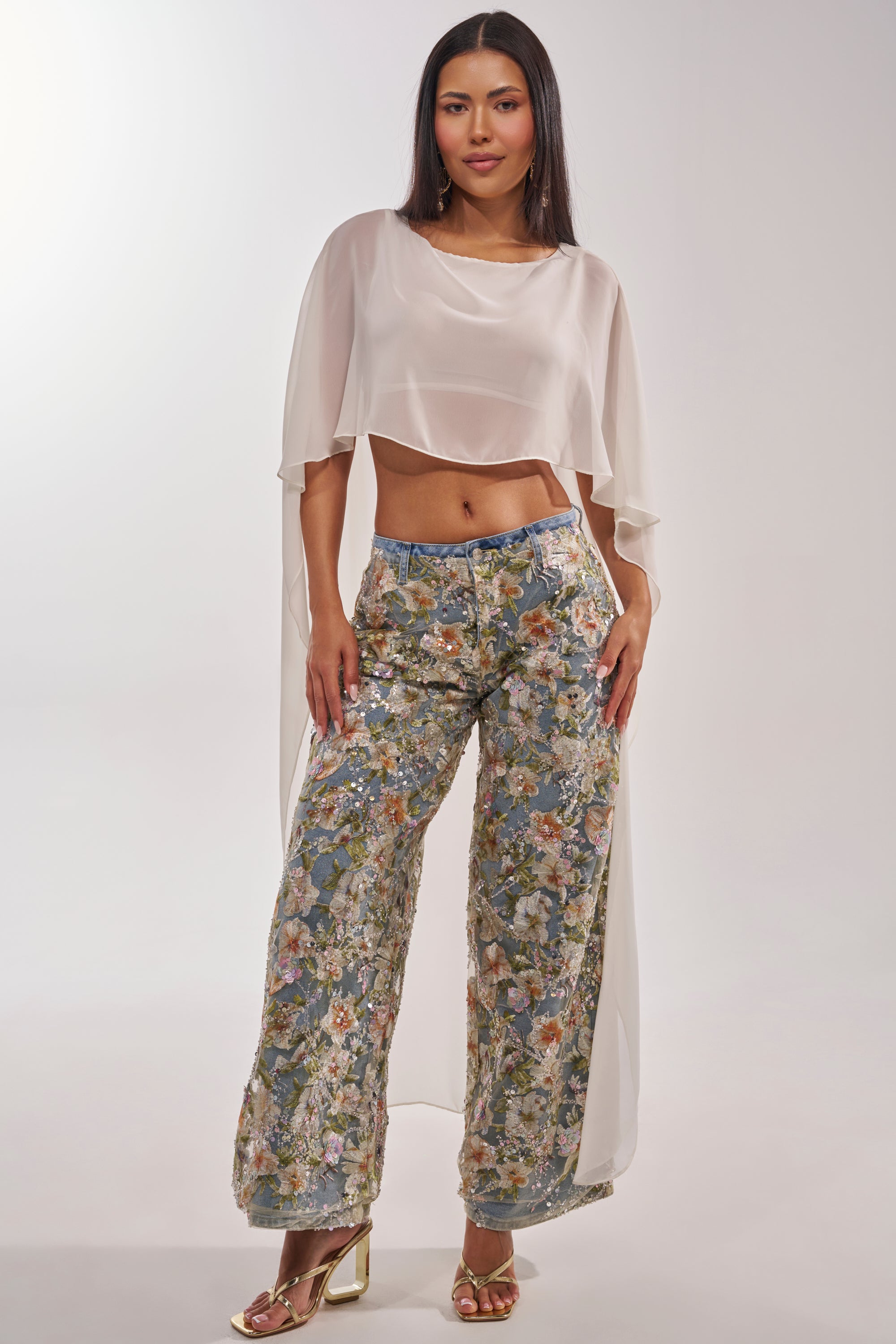 A woman poses confidently against a light background, wearing the IRIS RELAXED DENIM WITH FLORAL OVERLAY paired with a sheer white cape-style crop top and gold heels. 7