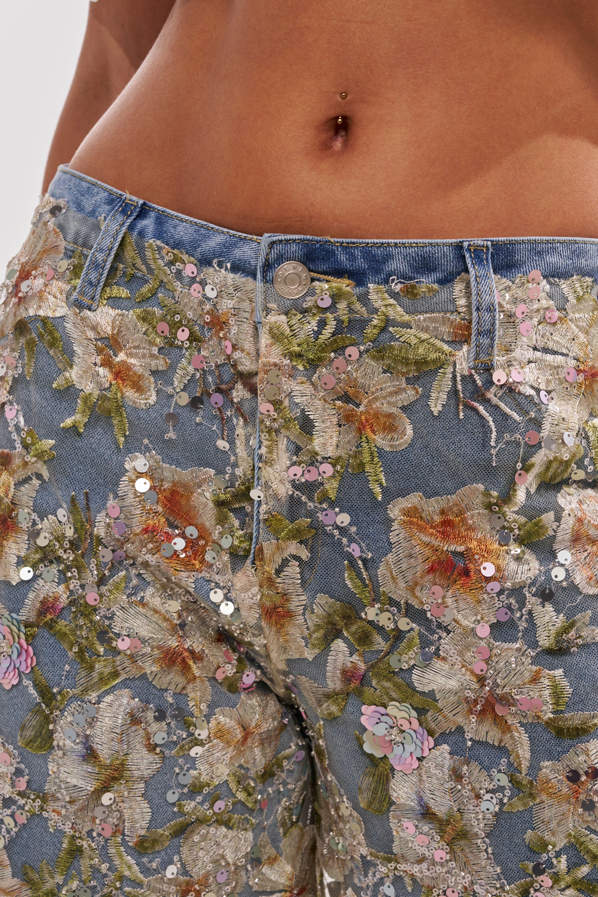 A close-up of a person wearing the IRIS RELAXED DENIM WITH FLORAL OVERLAY, featuring colorful floral embroidery and sequins; their midriff is shown with a pierced belly button. 8
