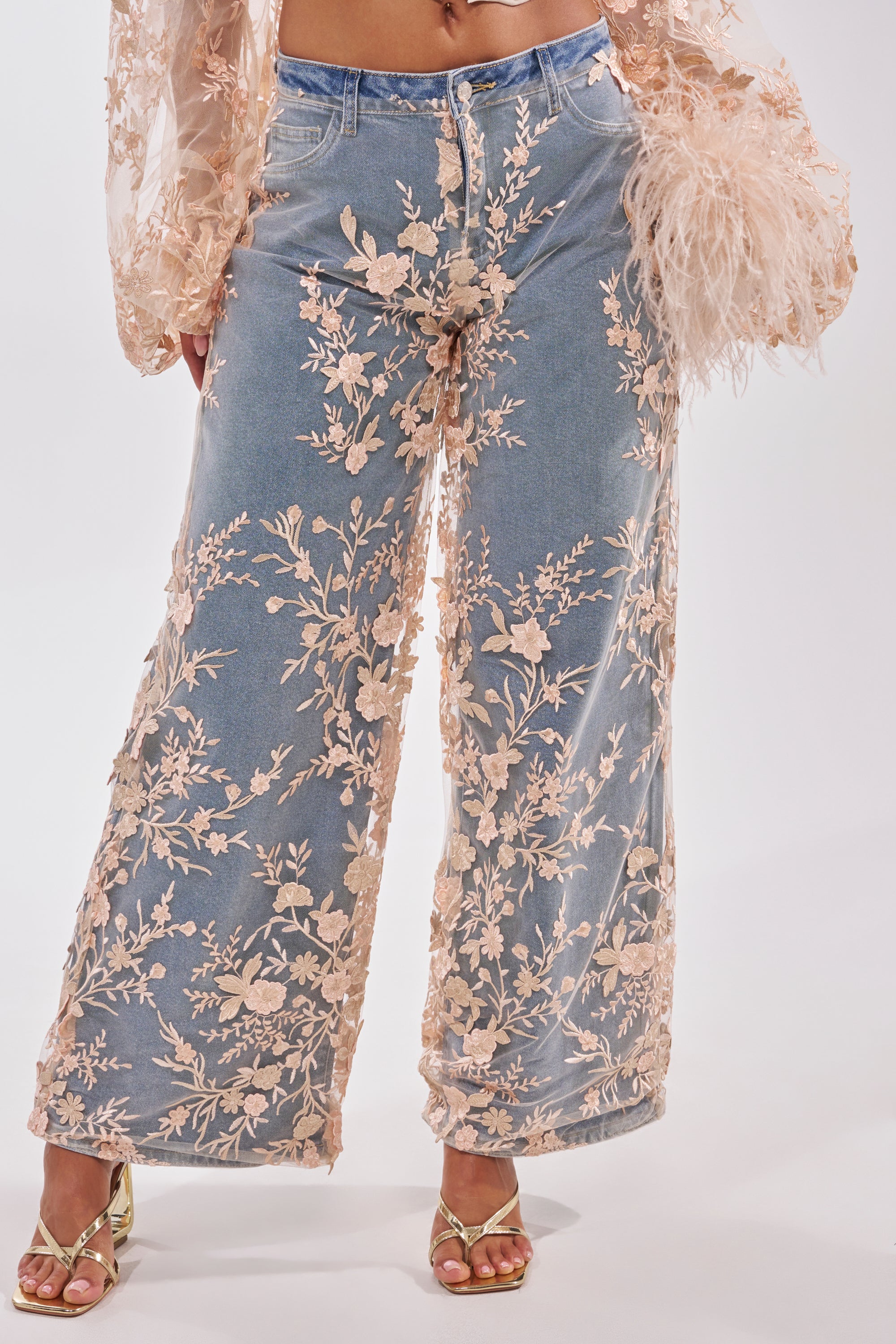 Only the lower half of the body is visible, showing FLY AWAY WITH ME FLORAL DENIM: high-rise, wide-leg jeans with intricate pink floral embroidery, paired with a sheer feathered top and gold open-toe heels. 1