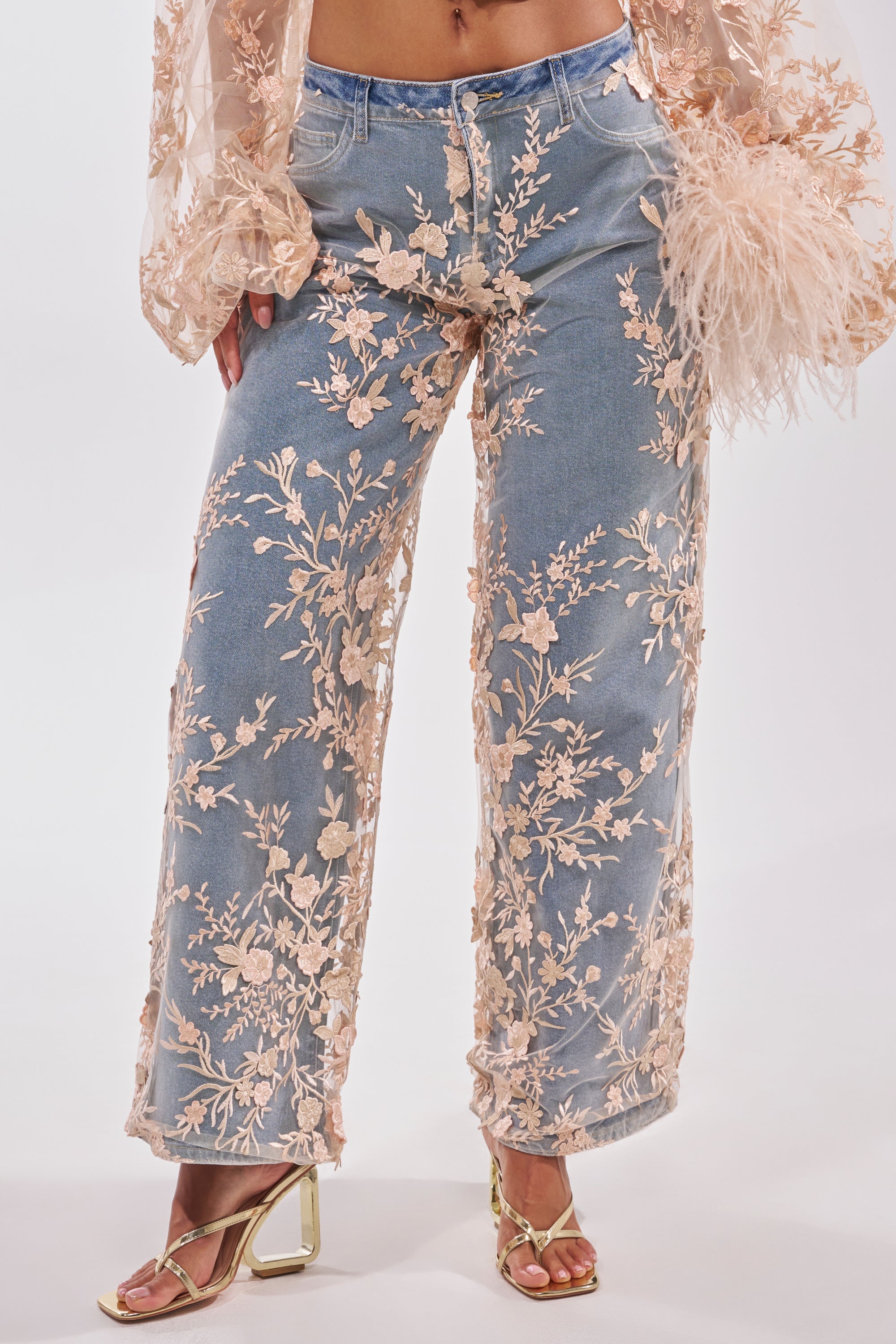 A person in the FLY AWAY WITH ME FLORAL DENIM: light blue, high-rise wide-leg jeans featuring sheer floral embroidery, styled with a matching sheer blouse with feathered cuffs and gold open-toe heeled sandals. 2
