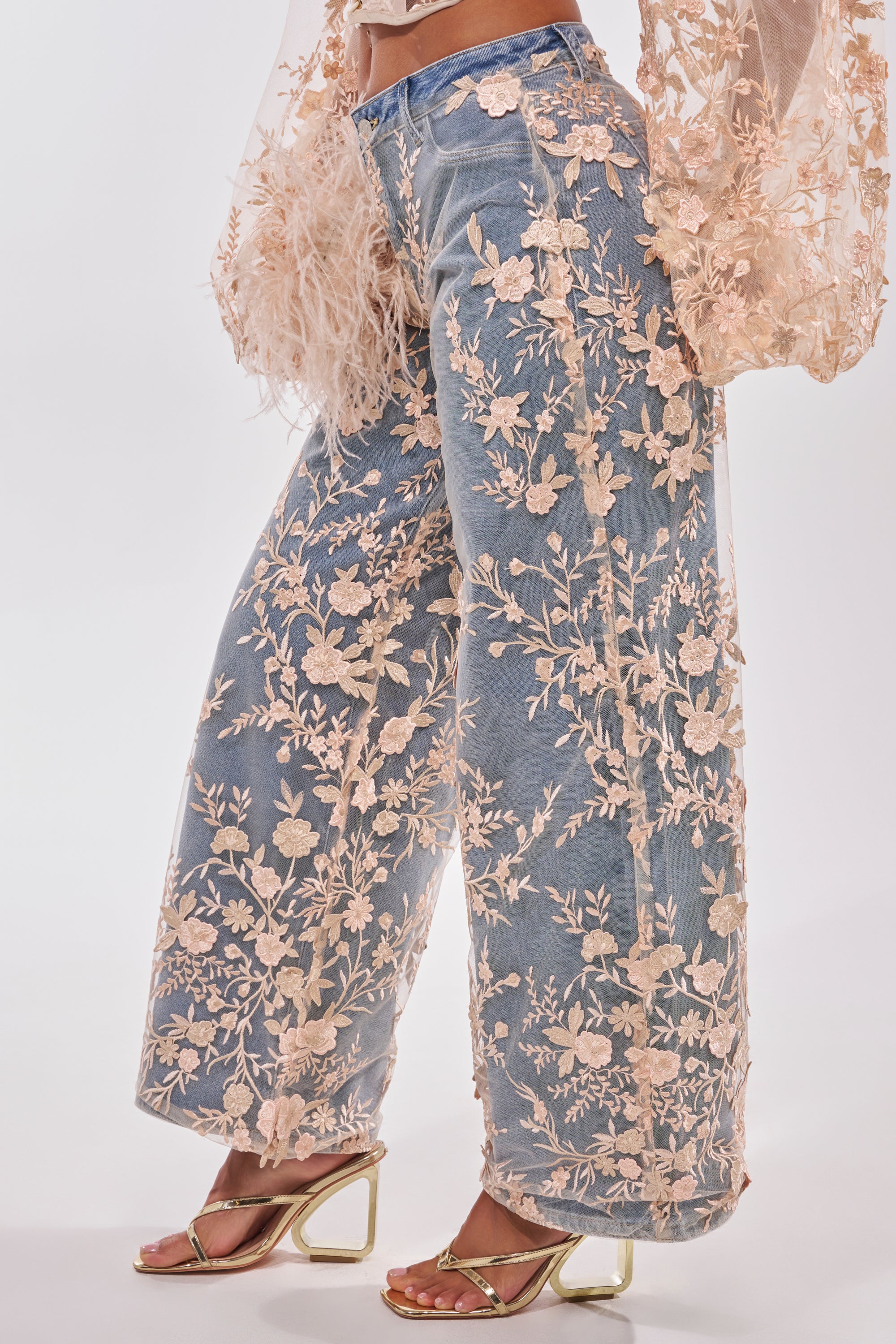 Wearing the FLY AWAY WITH ME FLORAL DENIM: wide-leg, high-rise blue jeans with sheer peach floral mesh overlay, paired with a matching embroidered mesh top and gold open-toe heels. 3