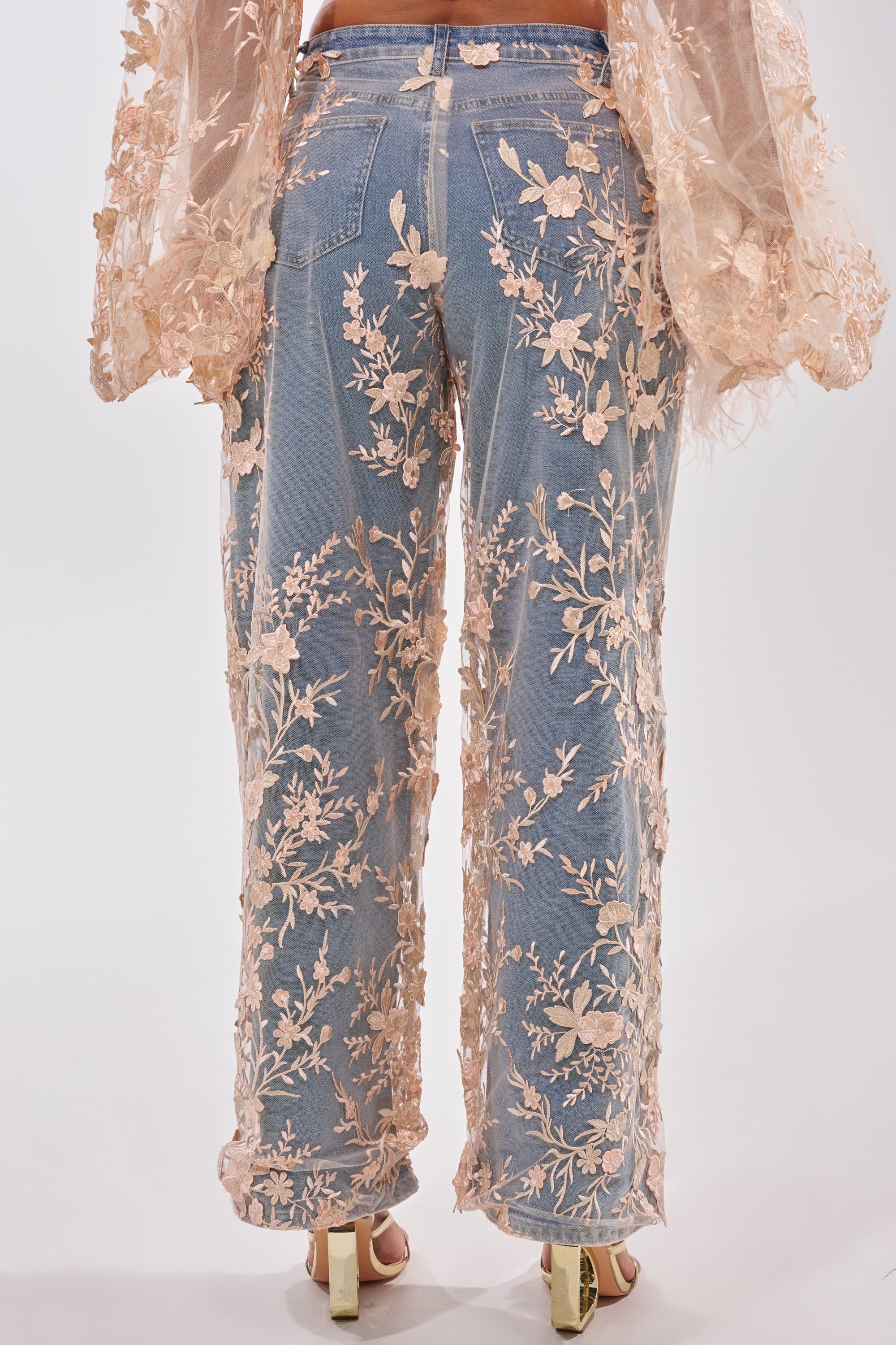A person wears the FLY AWAY WITH ME FLORAL DENIM set—wide-leg blue jeans and a matching sheer floral mesh overlay top—viewed from the back, styled with open-toe heels against a plain background. 4