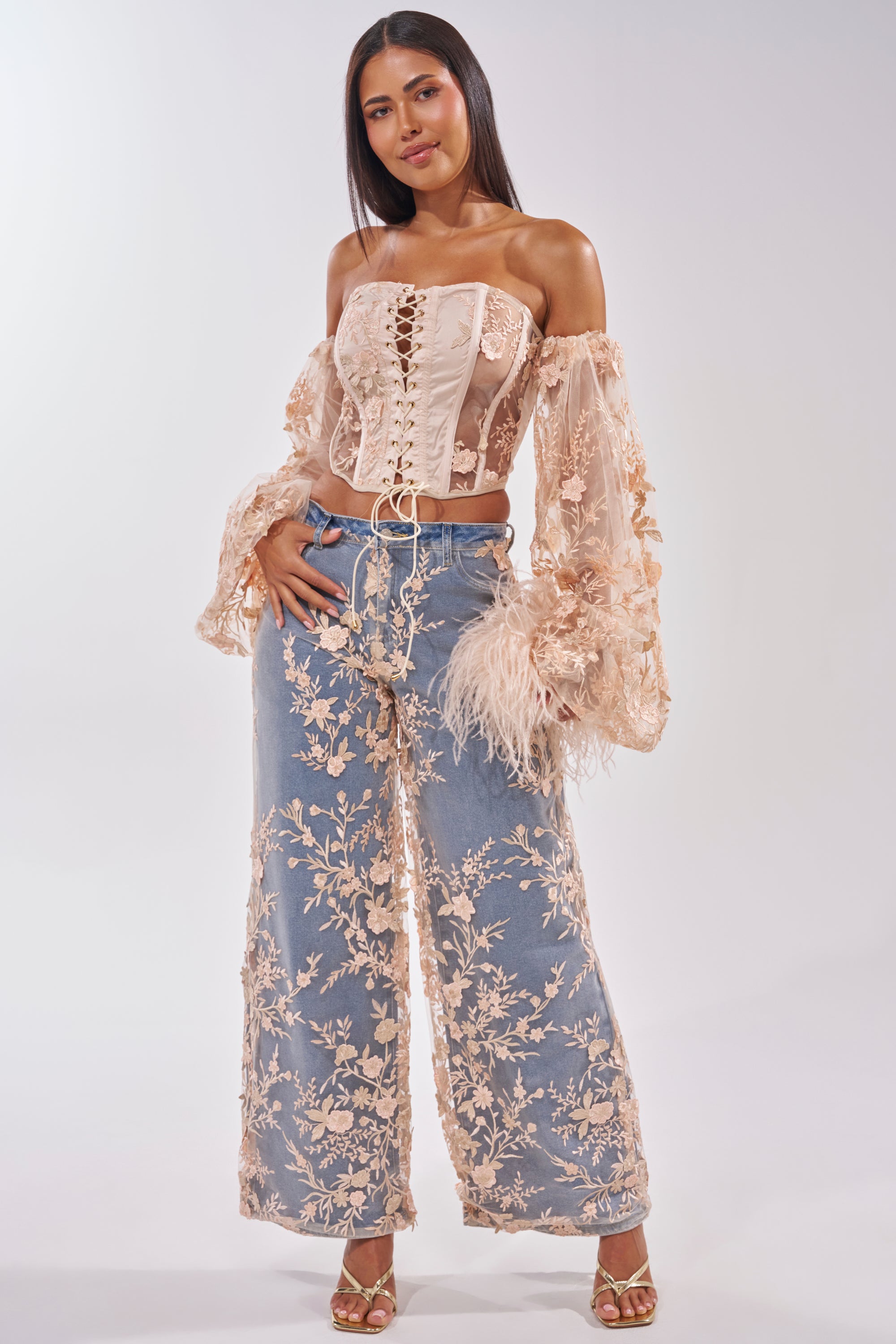 A woman models the FLY AWAY WITH ME FLORAL DENIM—high-rise, wide-leg jeans with a floral mesh overlay—styled with a sheer off-the-shoulder lace-up corset top and strappy heels against a light background. 5