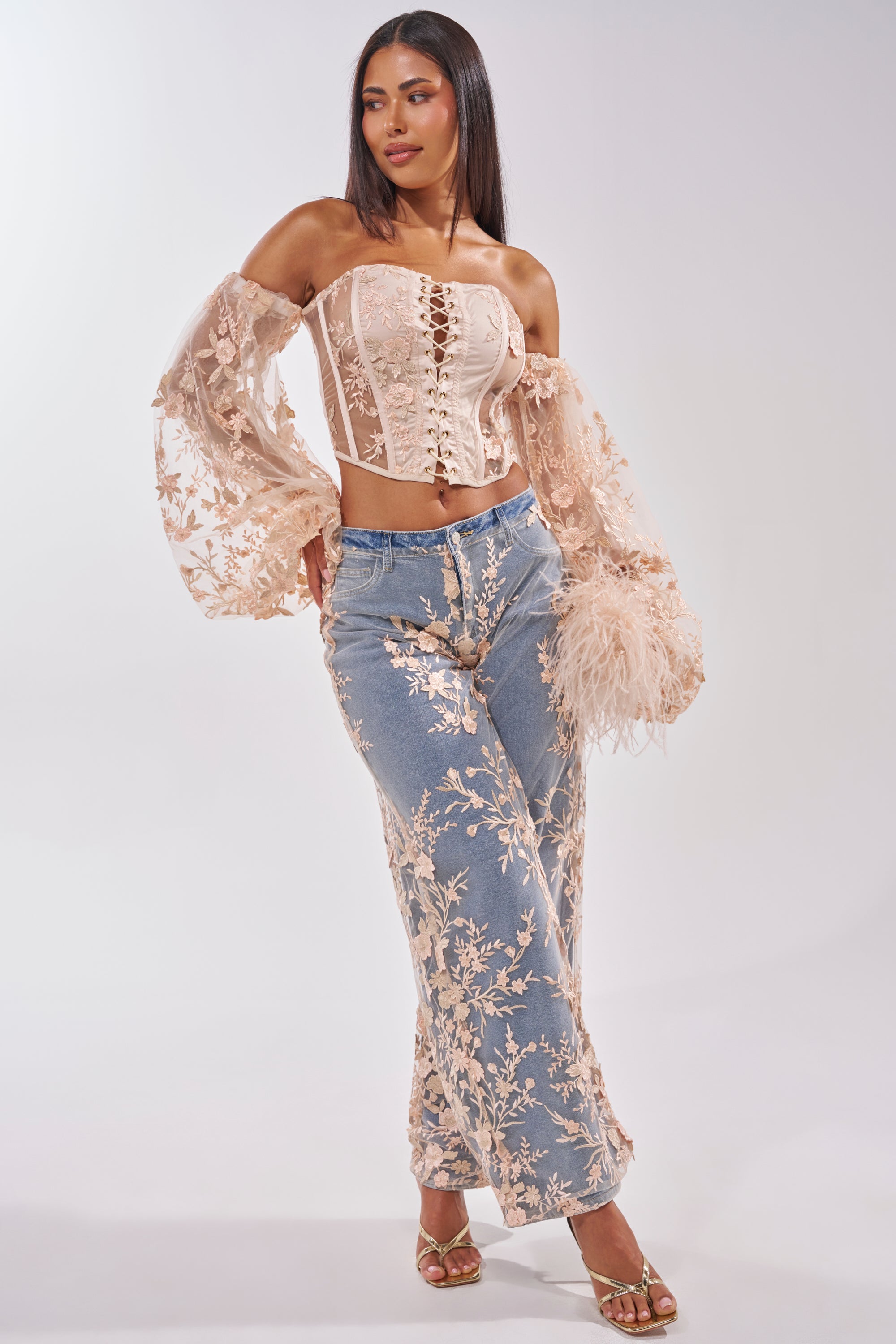 A woman stands confidently in the FLY AWAY WITH ME FLORAL DENIM—blue wide-leg jeans with a high-rise waist and pink floral embroidery—styled with a blush pink lace corset top, sheer puffy sleeves, and gold sandals against a plain backdrop. 7