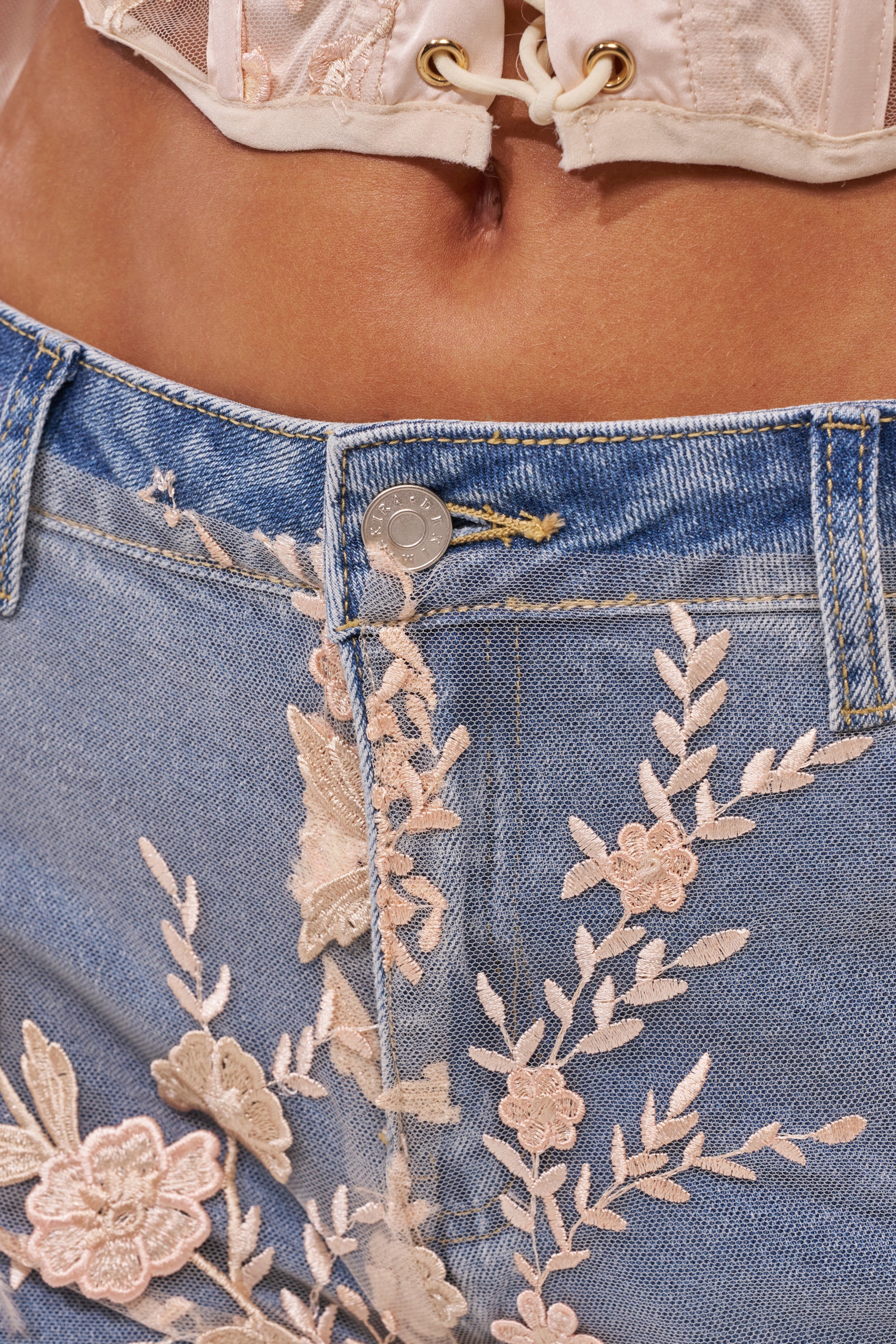 Close-up of a person wearing the FLY AWAY WITH ME FLORAL DENIM high-rise light blue jeans with pink floral embroidery and a cream corset top, photo cropped to display the waist and part of the torso. 8