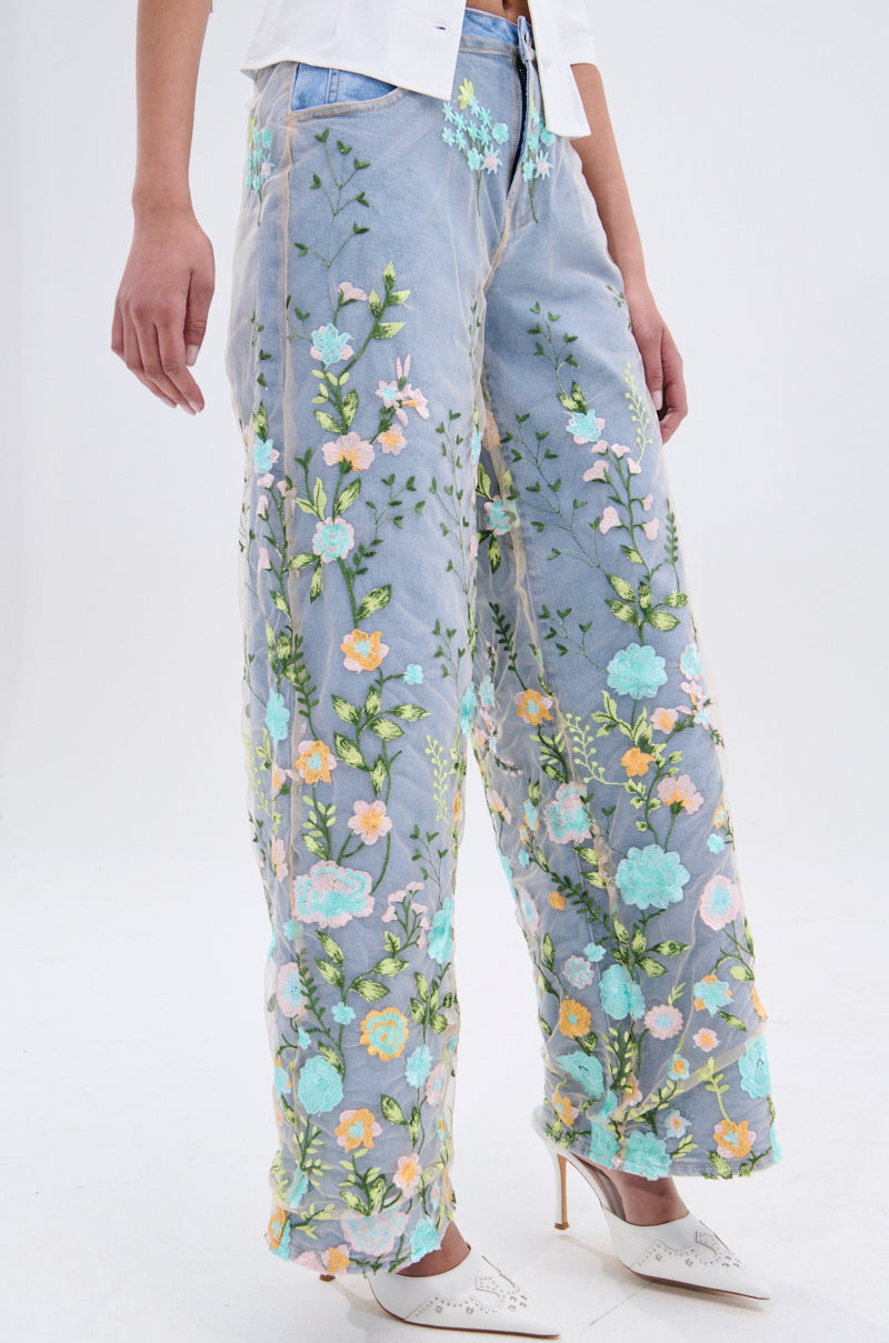 Wearing the FLOWER POWER RELAXED DENIM, featuring wide-leg light blue denim with colorful floral embroidery, paired with white pointed-toe heels and a white top.