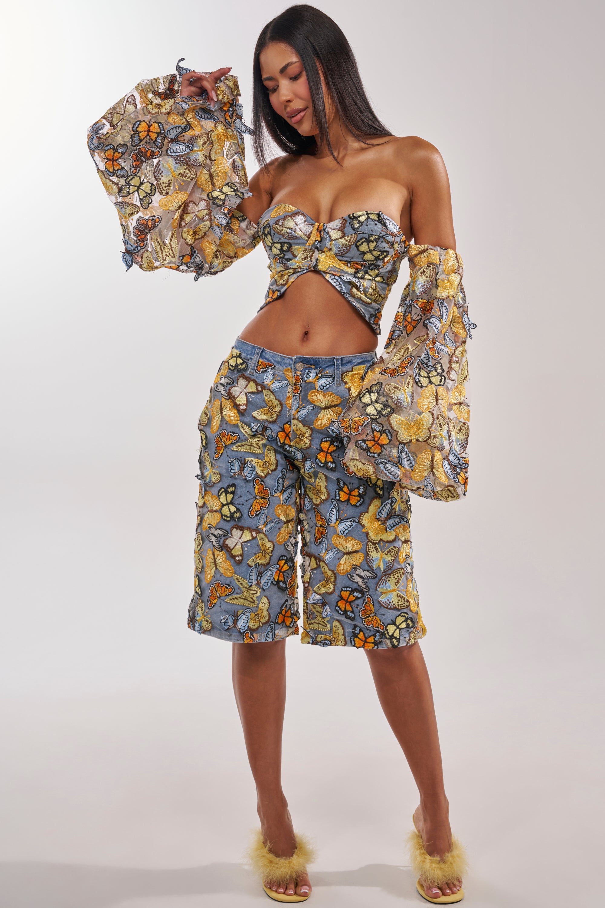 A woman wears the BUTTERFLY EFFECT BERMUDA SHORT with a blue and yellow butterfly print, paired with a strapless crop top, sheer off-the-shoulder sleeves, and fluffy yellow heels—ideal for festivals or standout street style. 3