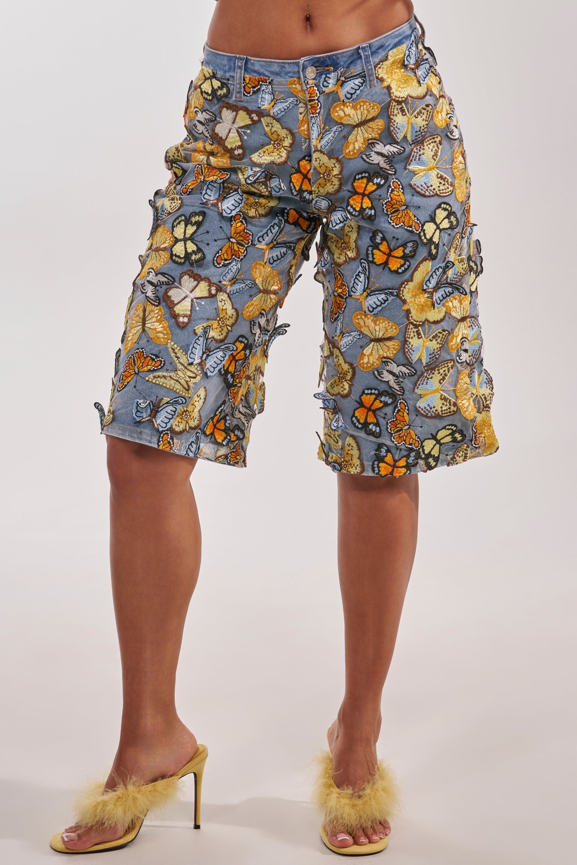 A person in statement streetwear: BUTTERFLY EFFECT BERMUDA SHORTS and yellow fuzzy high-heeled sandals, standing against a plain background. 4