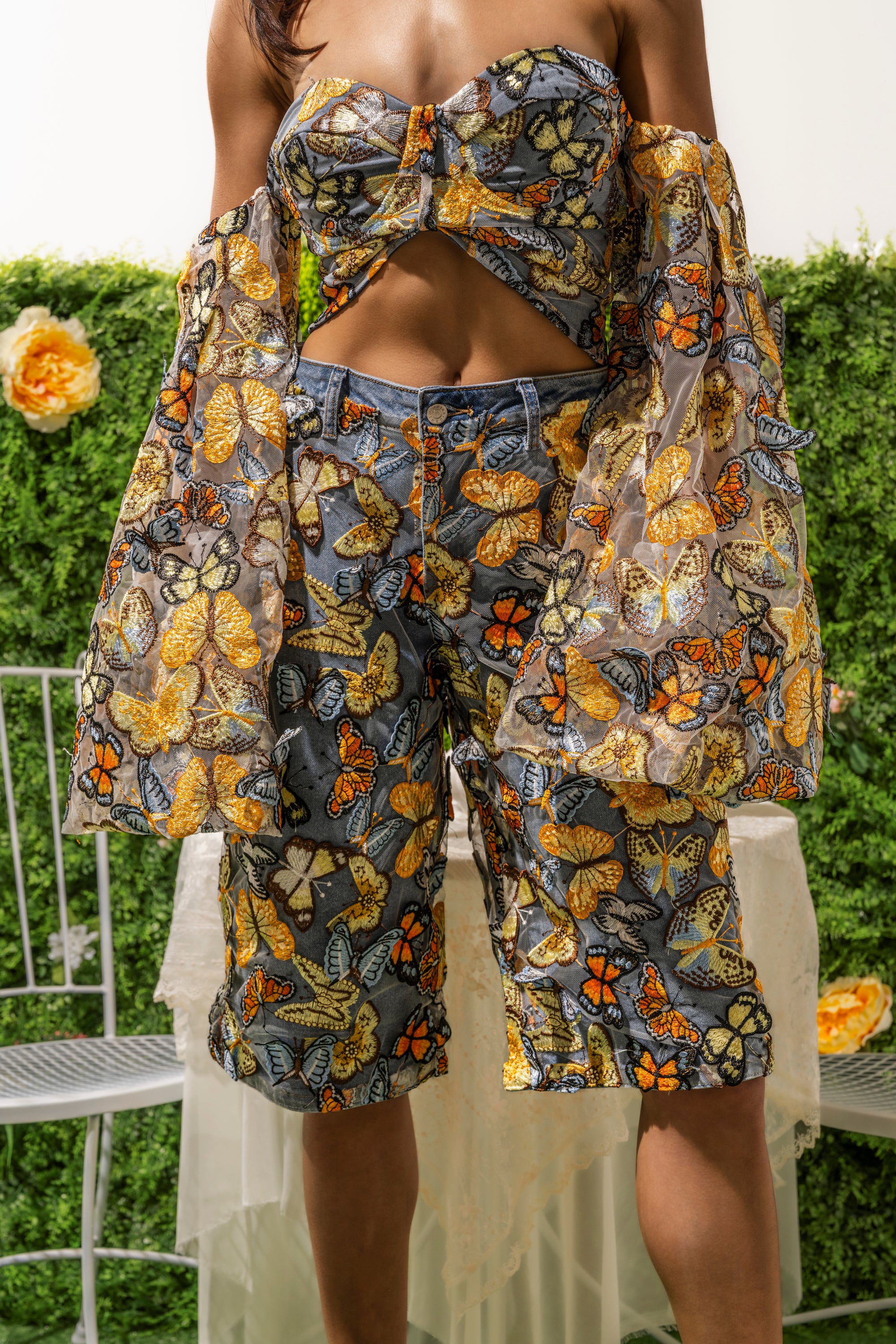A person wearing a butterfly-patterned, off-shoulder top and statement BUTTERFLY EFFECT BERMUDA SHORT stands in front of a green, flower-decorated hedge and a silver chair. 1
