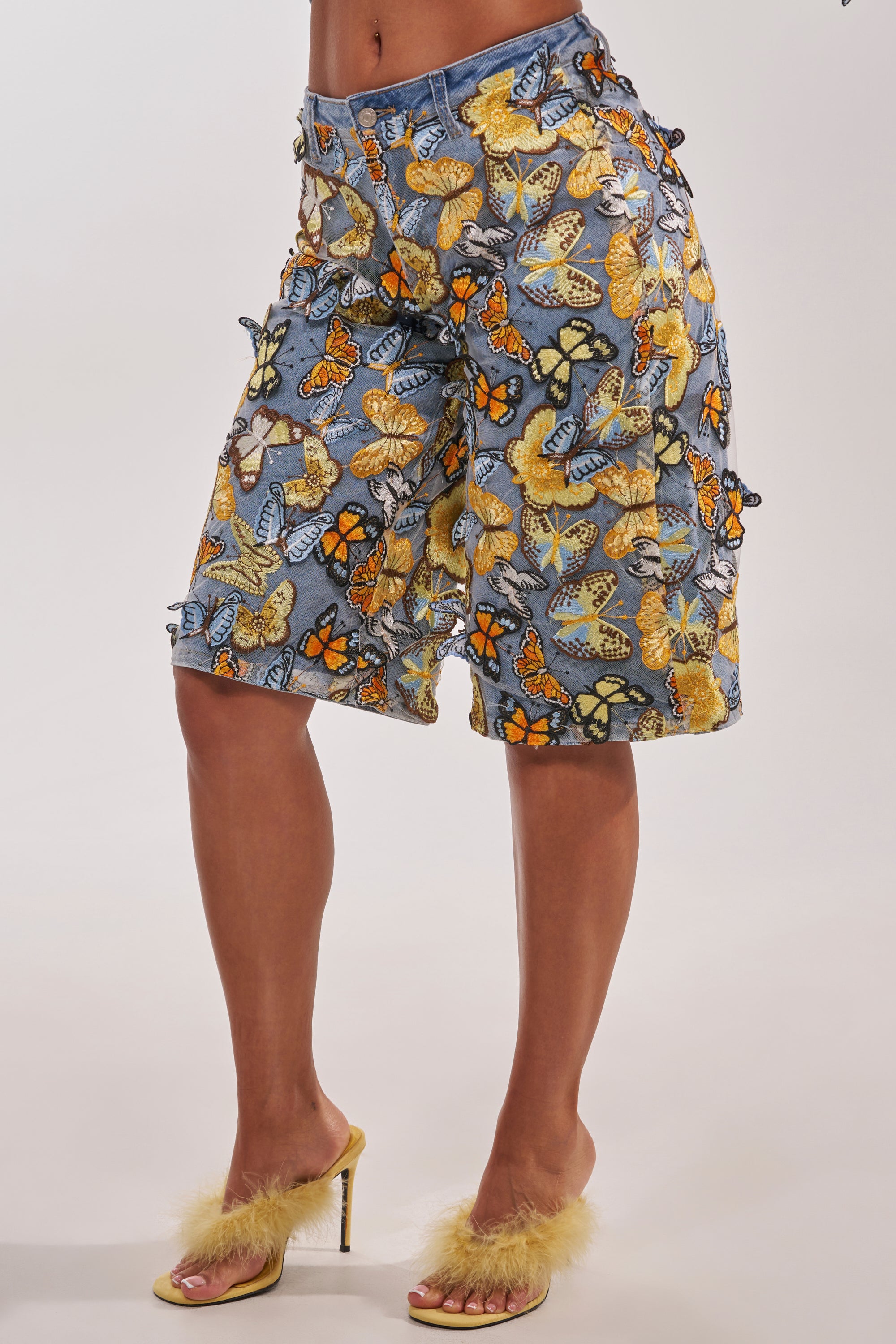Wearing the BUTTERFLY EFFECT BERMUDA SHORT and yellow high-heeled sandals with fluffy details, a person poses confidently against a plain background, showcasing bold statement streetwear style. 5