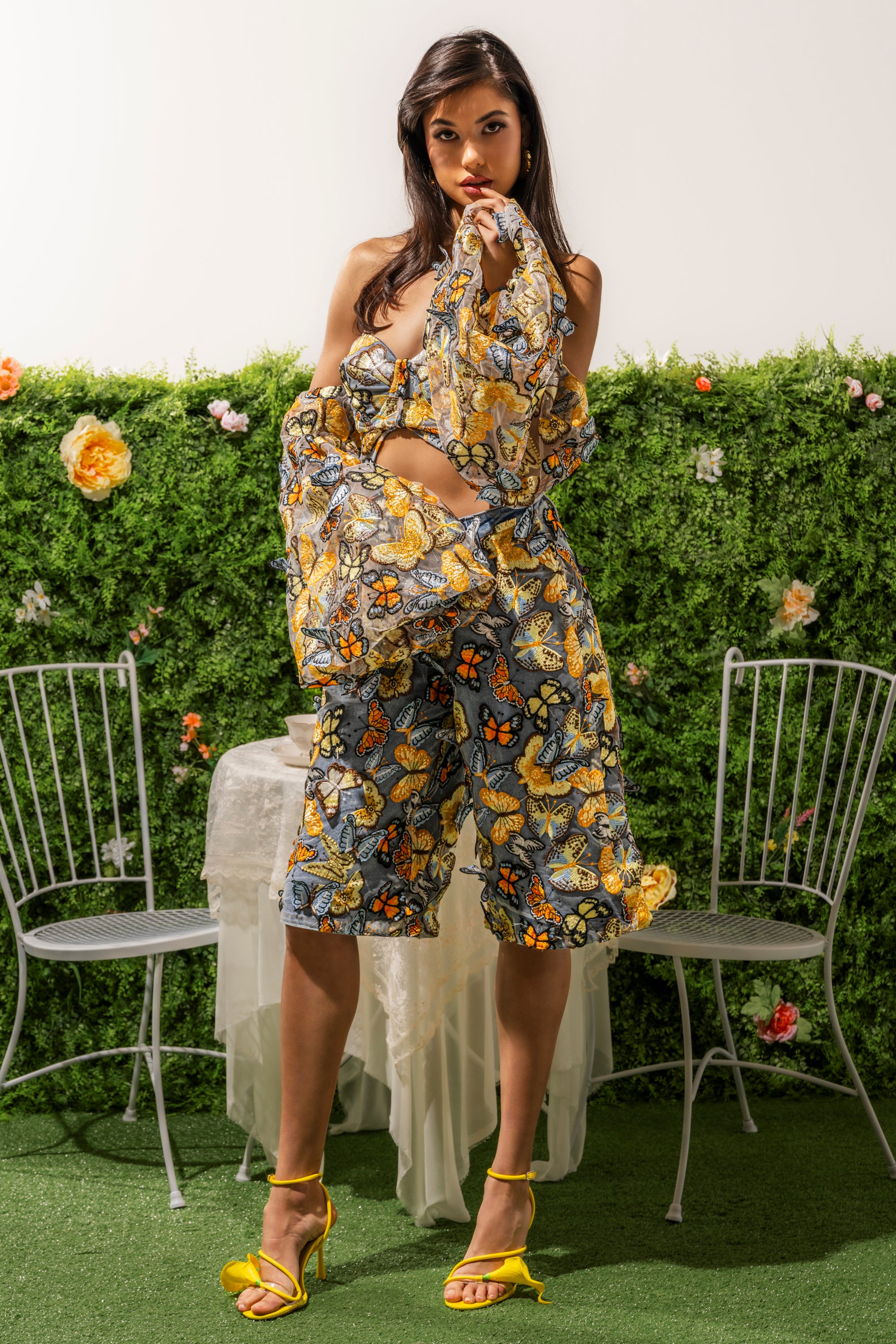 A woman poses outdoors in the BUTTERFLY EFFECT BERMUDA SHORT and a vibrant butterfly-print top. She stands between metal chairs and a table, with yellow heels, flowers, and greenery completing her bold streetwear look. 2