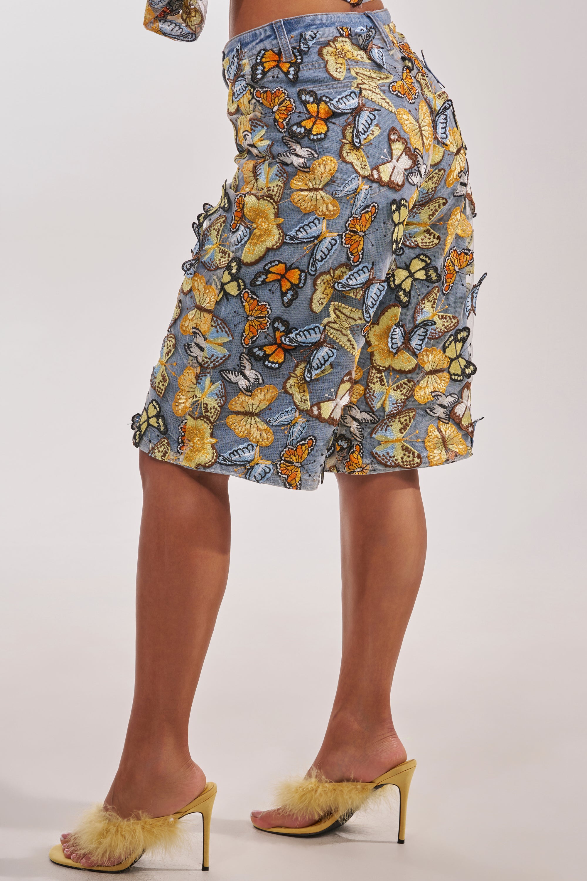 Wearing the BUTTERFLY EFFECT BERMUDA SHORT, featuring blue denim with colorful embroidered butterflies, and yellow-heeled sandals with fluffy trim, a person channels playful streetwear style against a plain light background. 6