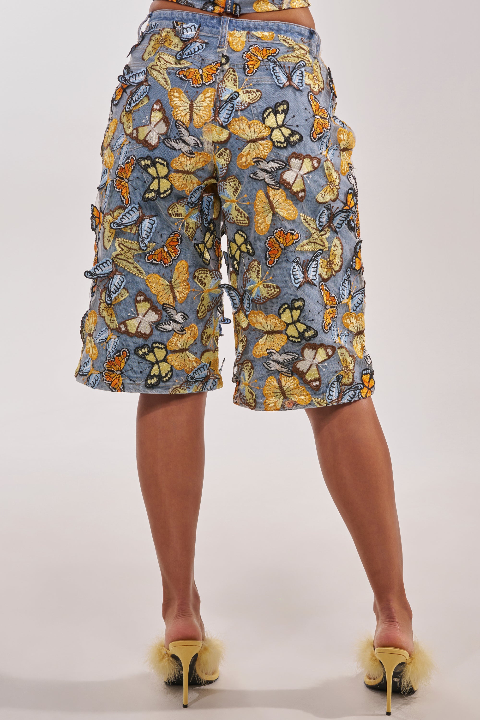 Person standing with their back to the camera, wearing the BUTTERFLY EFFECT BERMUDA SHORT covered in colorful patterns and yellow fluffy-accented heels—a bold choice for standout festival outfits. 7