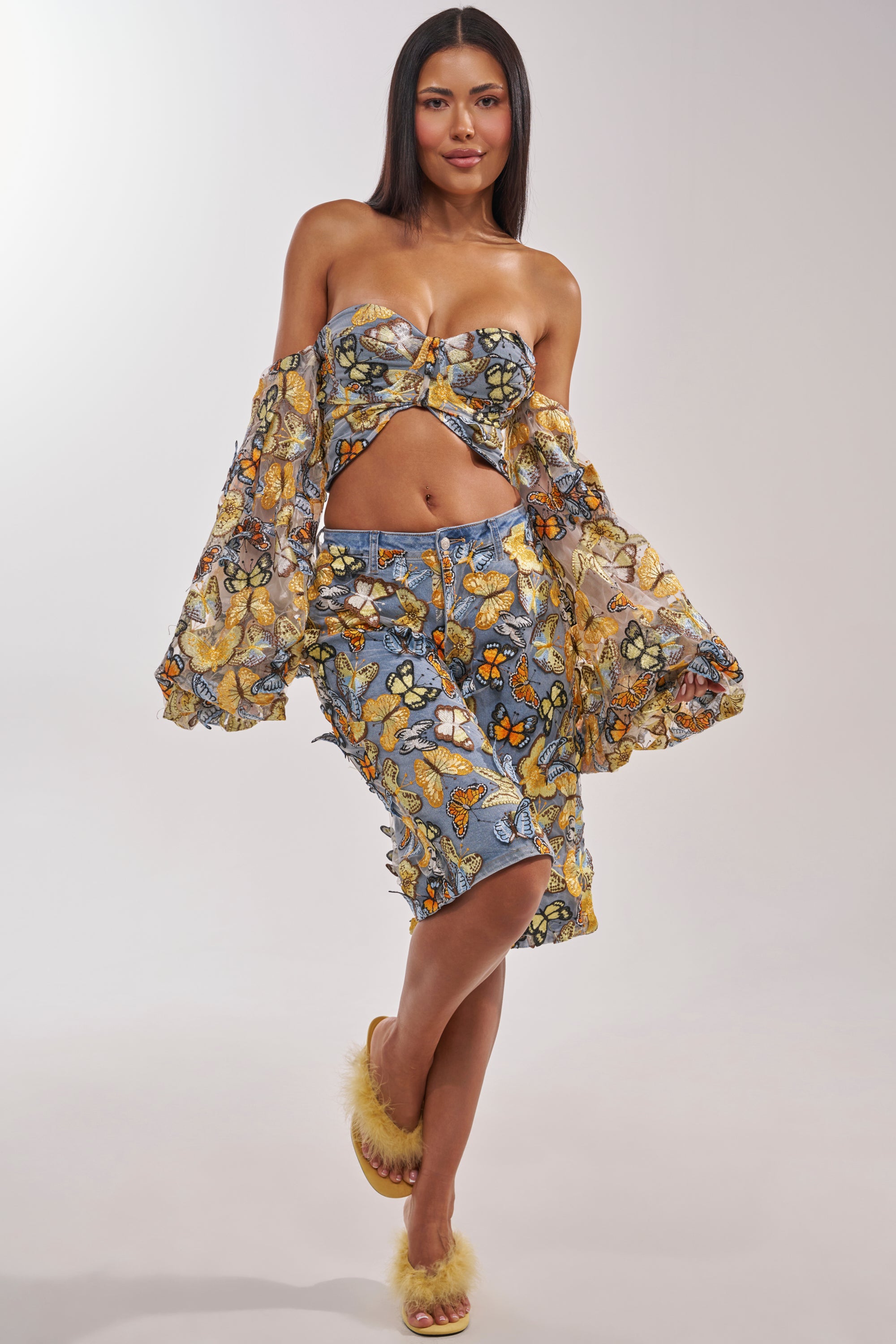 A woman poses confidently in BUTTERFLY EFFECT BERMUDA SHORTS with a matching floral top and flowing sheer sleeve—ideal for festival outfits. She wears fuzzy yellow slippers and has long, straight hair against a plain white background. 8