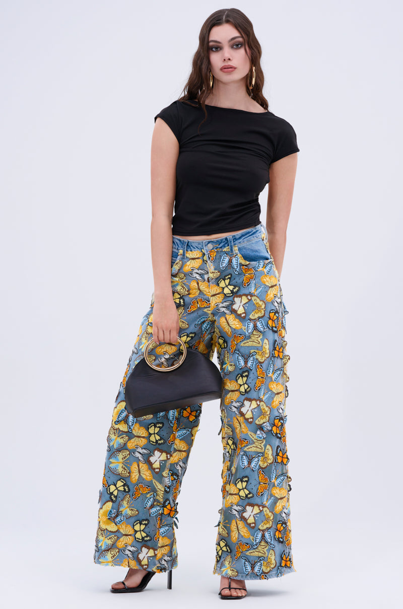 Wearing a black top, Butterfly Relaxed Denim with embroidered butterfly patches, and high-heeled shoes, someone holds a black handbag with circular handles. Their long wavy hair complements the plain, light background that highlights the colorful denim design.