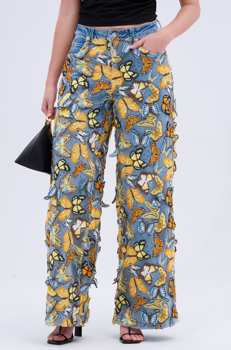 Against a plain background, a person in high-rise BUTTERFLY RELAXED DENIM with embroidered butterflies in orange, blue, and yellow holds a small black handbag and wears black heels.