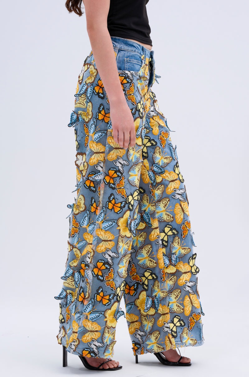 A person in Butterfly Relaxed Denim, featuring a colorful display of orange, yellow, and blue butterflies, paired with a black top and black heeled sandals against a plain light backdrop.
