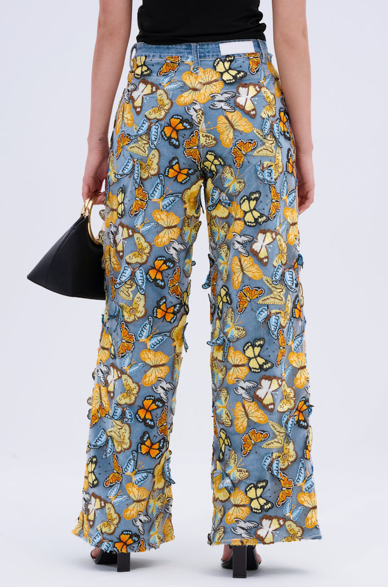A person wears BUTTERFLY RELAXED DENIM with vibrant embroidered butterfly patches in orange, yellow, and blue. They hold a black handbag and complete the outfit with sleek black shoes against a plain light gray background.