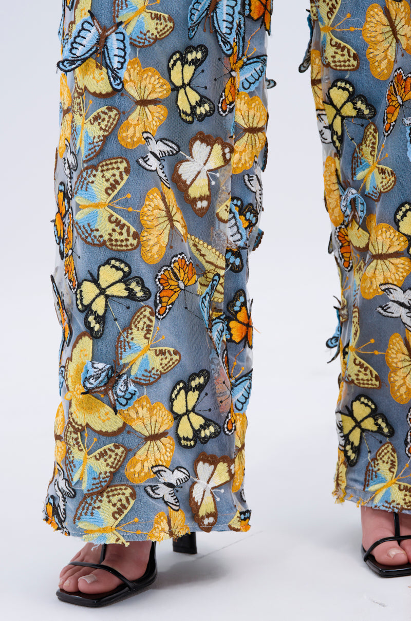 A person models Butterfly Relaxed Denim, featuring a vibrant pattern of yellow, blue, and orange butterflies on a gray background. The high-rise jeans are styled with black open-toed heeled sandals, blending style and charm effortlessly.