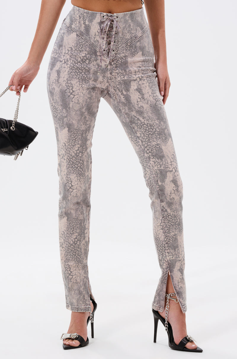 A person wearing IN THE SUN SNAKE LACE-UP PANT—a high-rise, lace-up, snakeskin-print pant with front ankle slits—black heeled sandals, and holding a small black chain-strap handbag. The background is plain white. 3