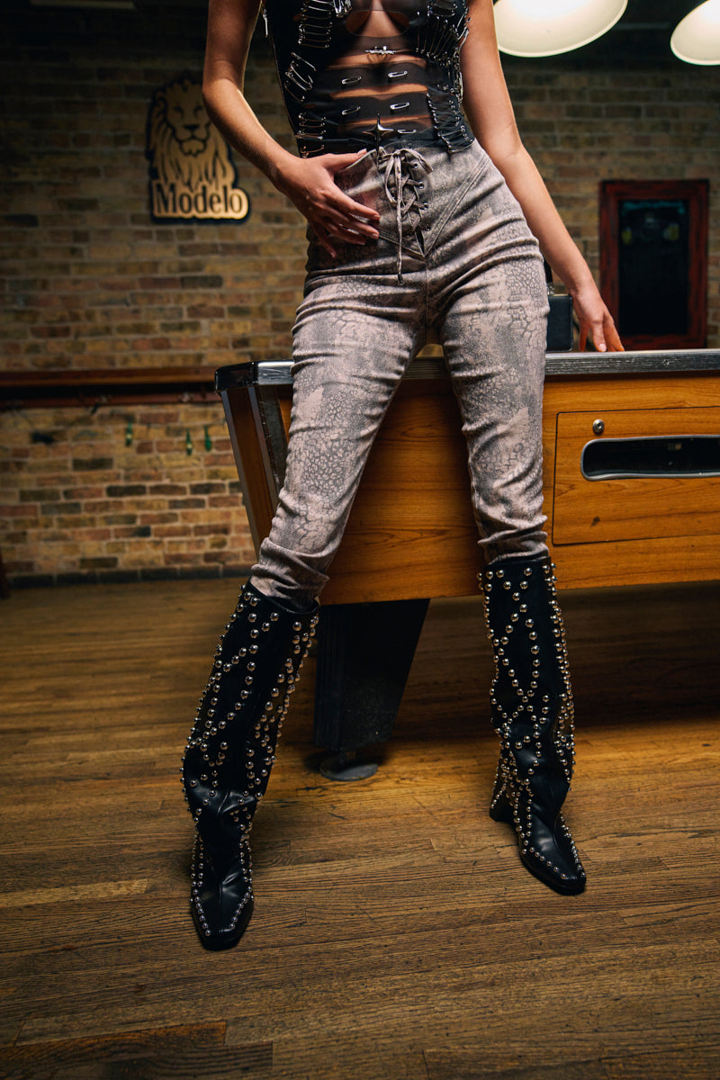 A person stands by a pool table wearing IN THE SUN SNAKE LACE-UP PANT, a black cutout top, and tall black boots with metallic chains and studs. Only the lower torso and legs are visible. A Modelo sign is on the brick wall behind. 2