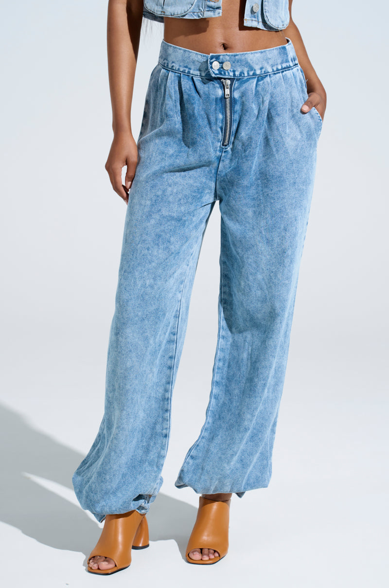 A person is wearing LEXI LIGHTWEIGHT DENIM PANT in light blue, featuring a high-waisted, relaxed silhouette with a visible zipper, matched with a similar top and complemented by tan open-toe heeled sandals. The background is plain and light-colored.