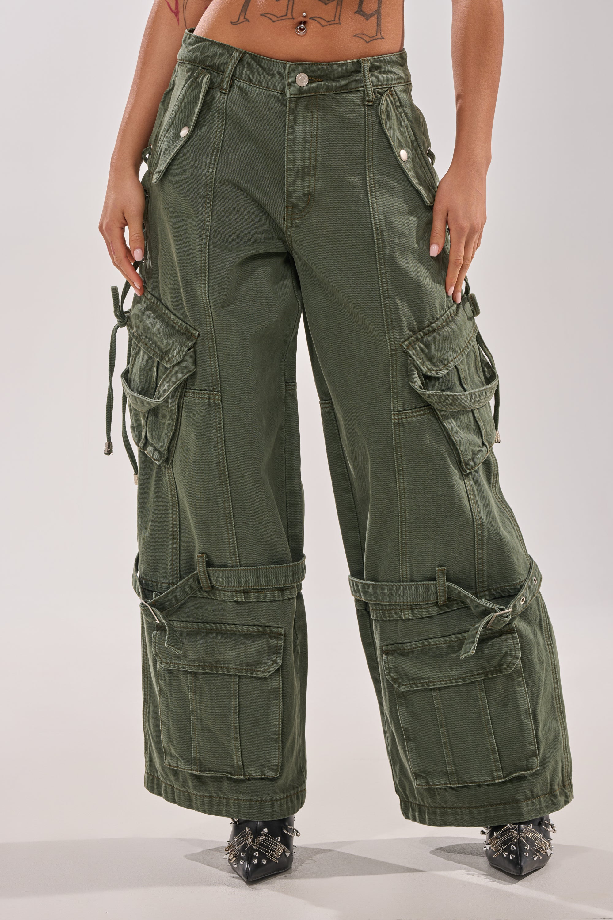 A person models the KARA CARGO DENIM—high-waisted, baggy olive pants with multiple large pockets, straps, and a wide-leg silhouette. Only their lower torso and legs are visible, hands relaxed at their sides. 1
