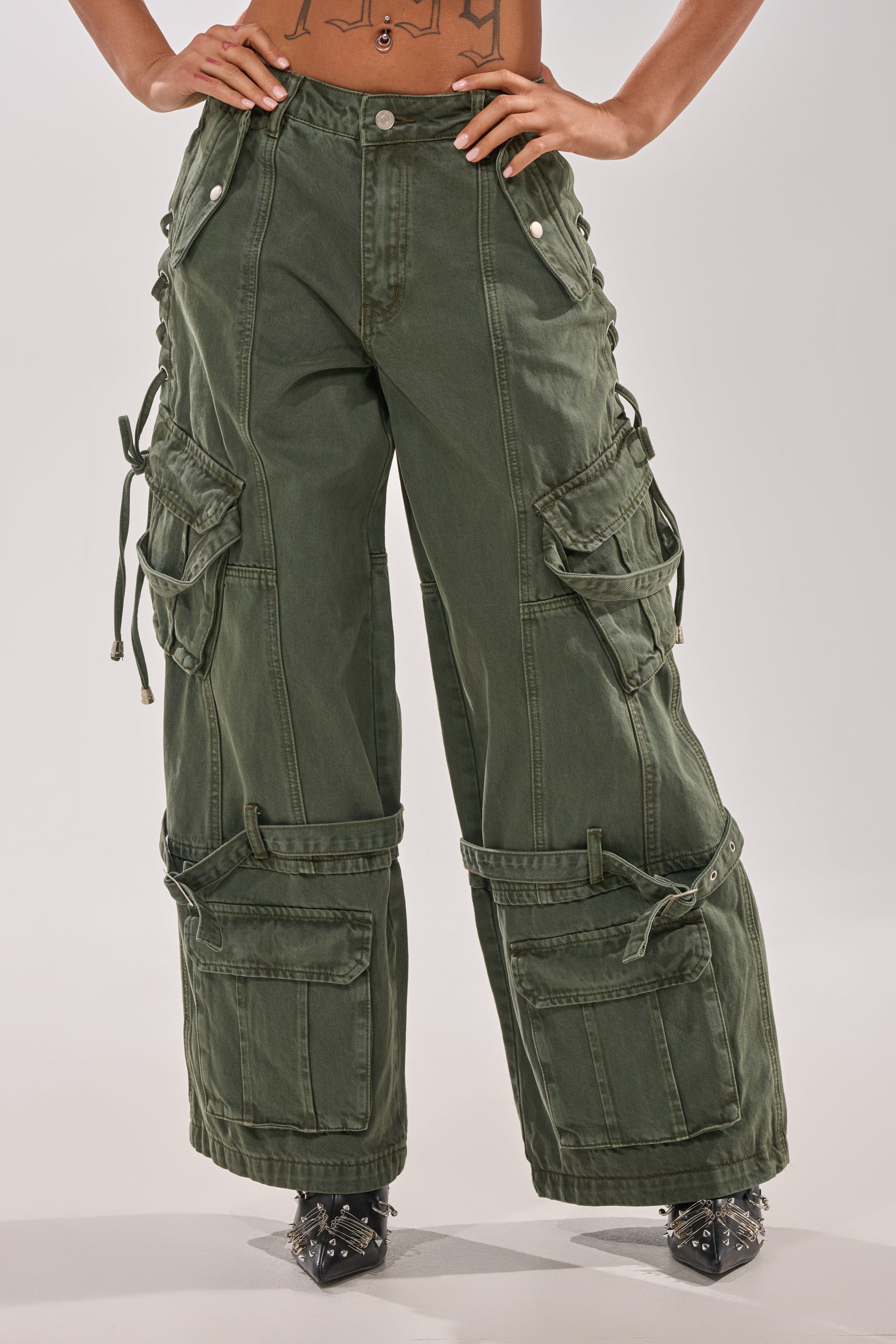Wearing KARA CARGO DENIM in olive, a high-waisted wide leg pant with multiple pockets, side straps, and button details. Person has hands on hips; pointed black shoes visible. Upper body is partially out of frame. 3