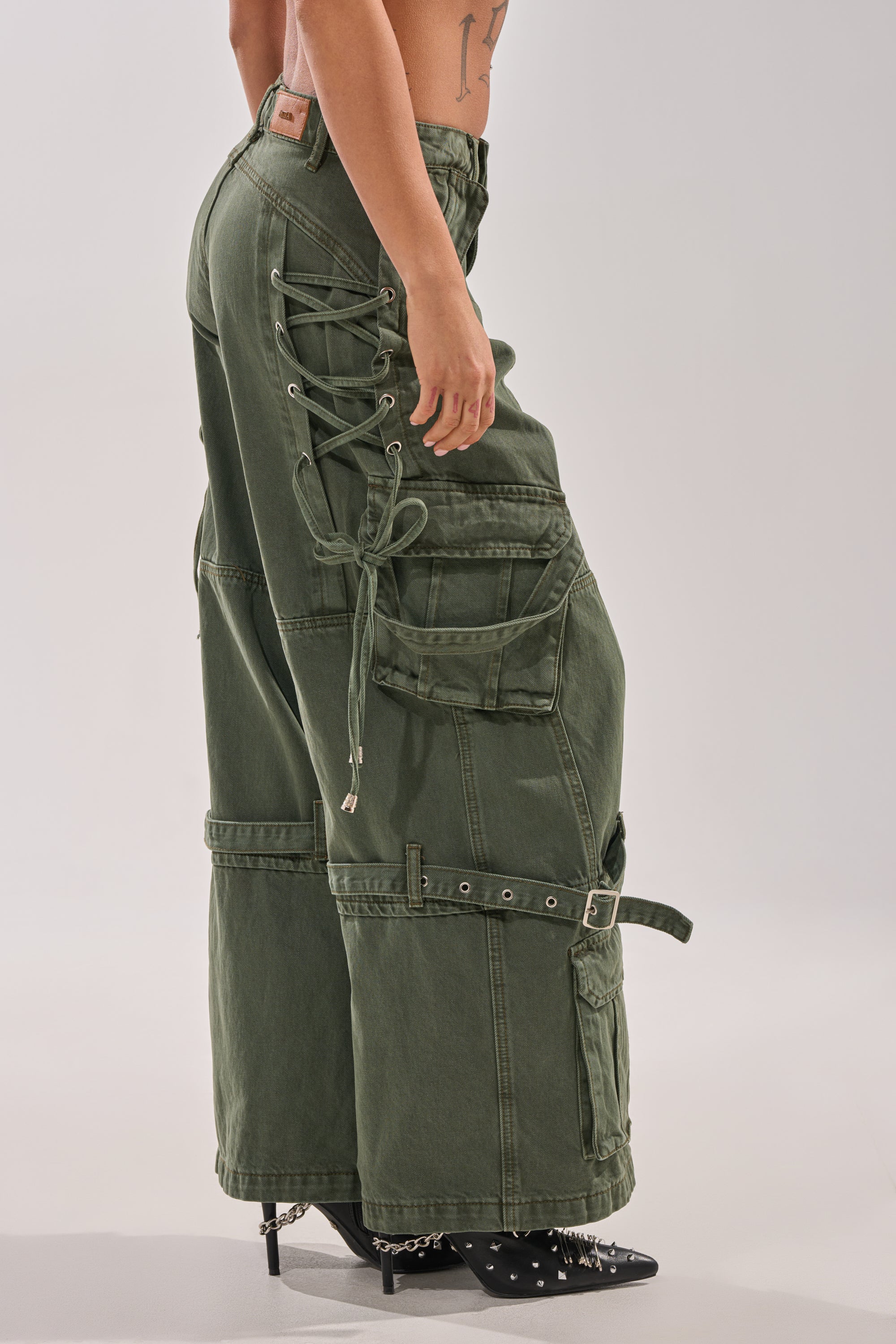 Wearing KARA CARGO DENIM—low-rise olive wide-leg pants with lace-up sides, multiple pockets, and a knee belt—paired with black pointed-toe shoes accented in silver. 4