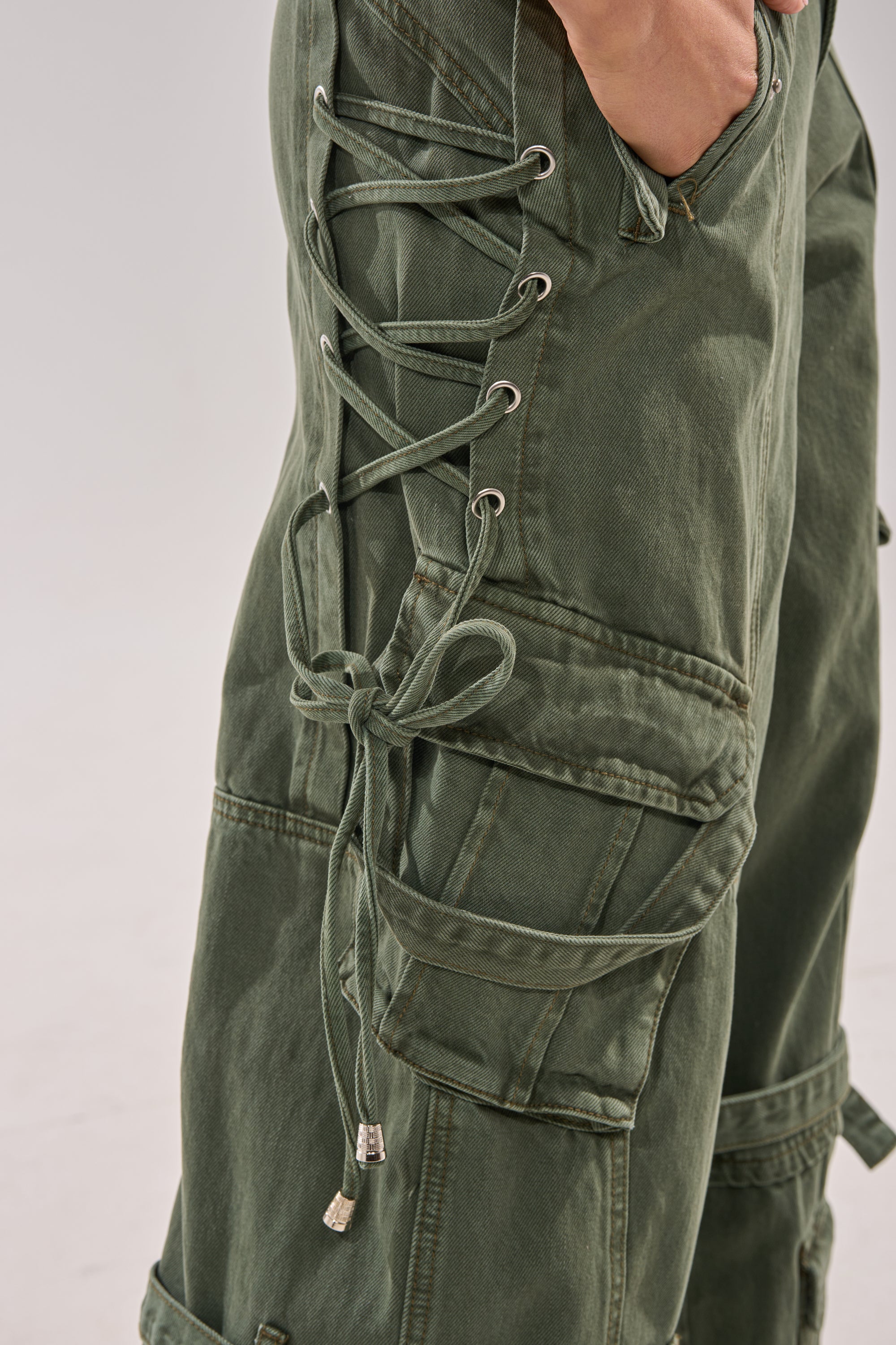 Close-up of KARA CARGO DENIM low-rise olive pants with lace-up and side bow details. Features a wide-leg silhouette, large cargo pockets, and a hand resting in the top pocket. 7