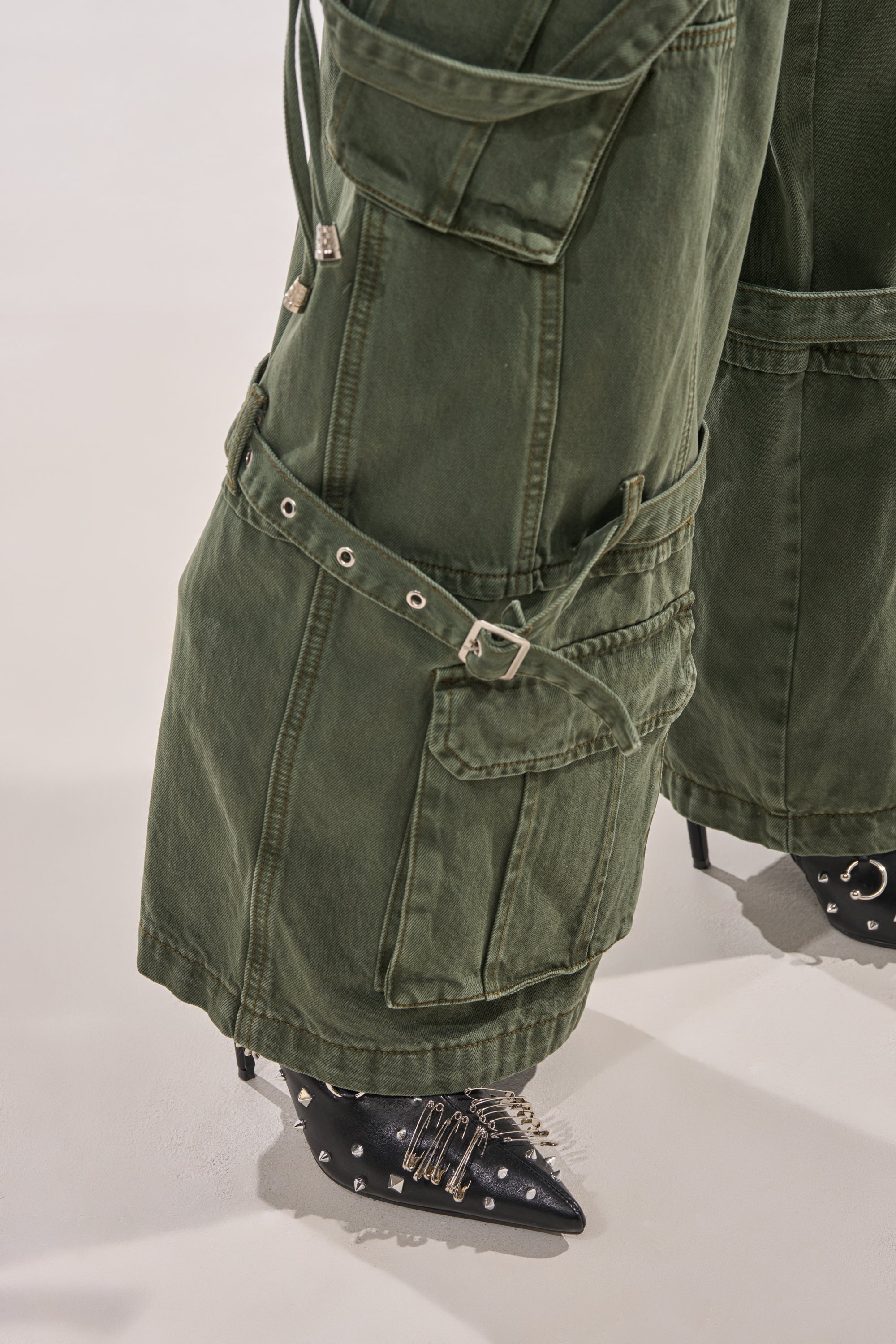 Close-up of the KARA CARGO DENIM pants in olive, featuring a wide leg, multiple pockets, straps, and buckles, styled with black pointed-toe shoes adorned with silver spikes and embellishments. 8