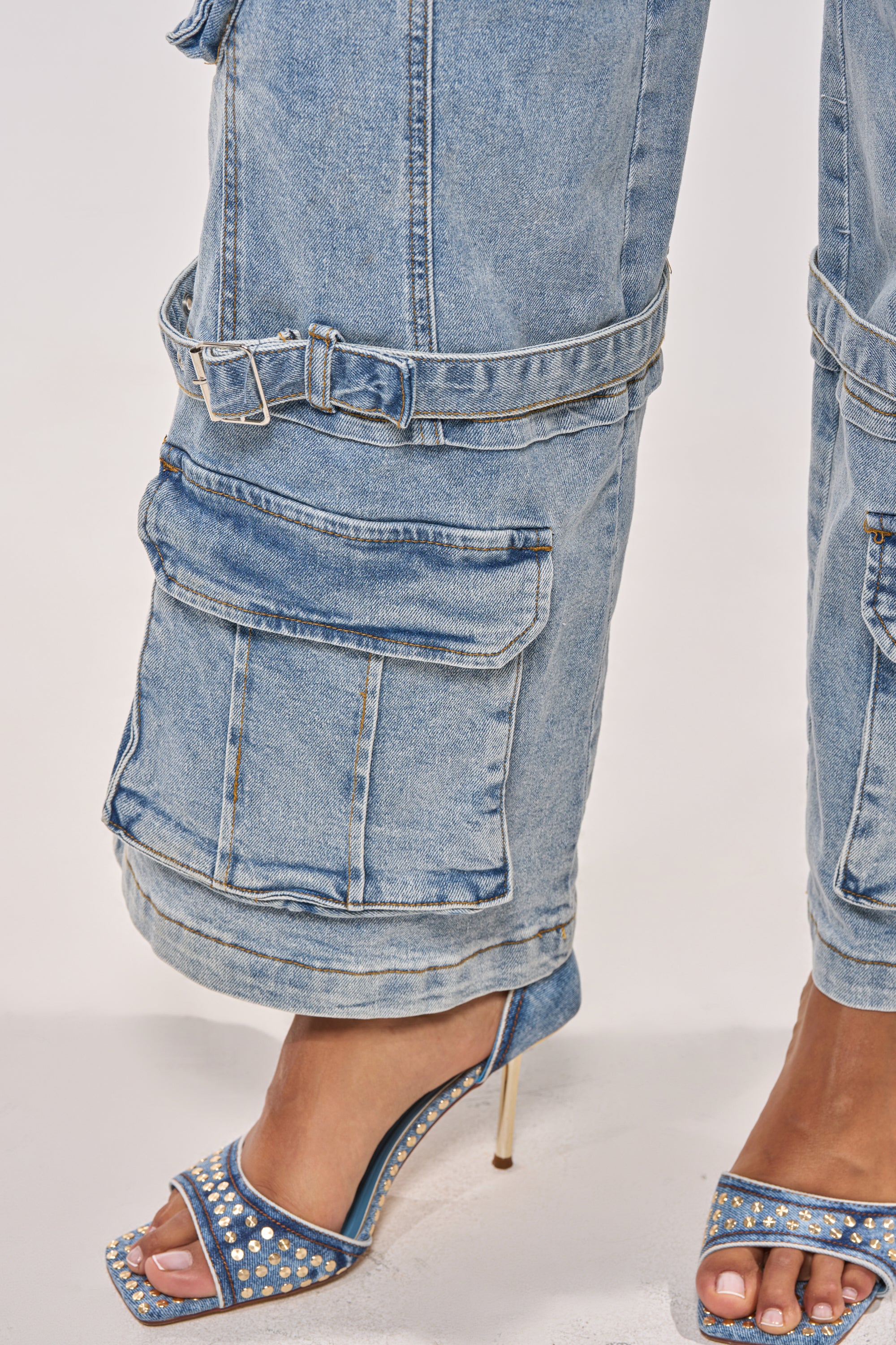 A close-up of a person wearing KARA DENIM CARGO PANT, featuring light wash denim, large pockets, and adjustable buckles, paired with blue studded high-heeled sandals against a plain light background. 12