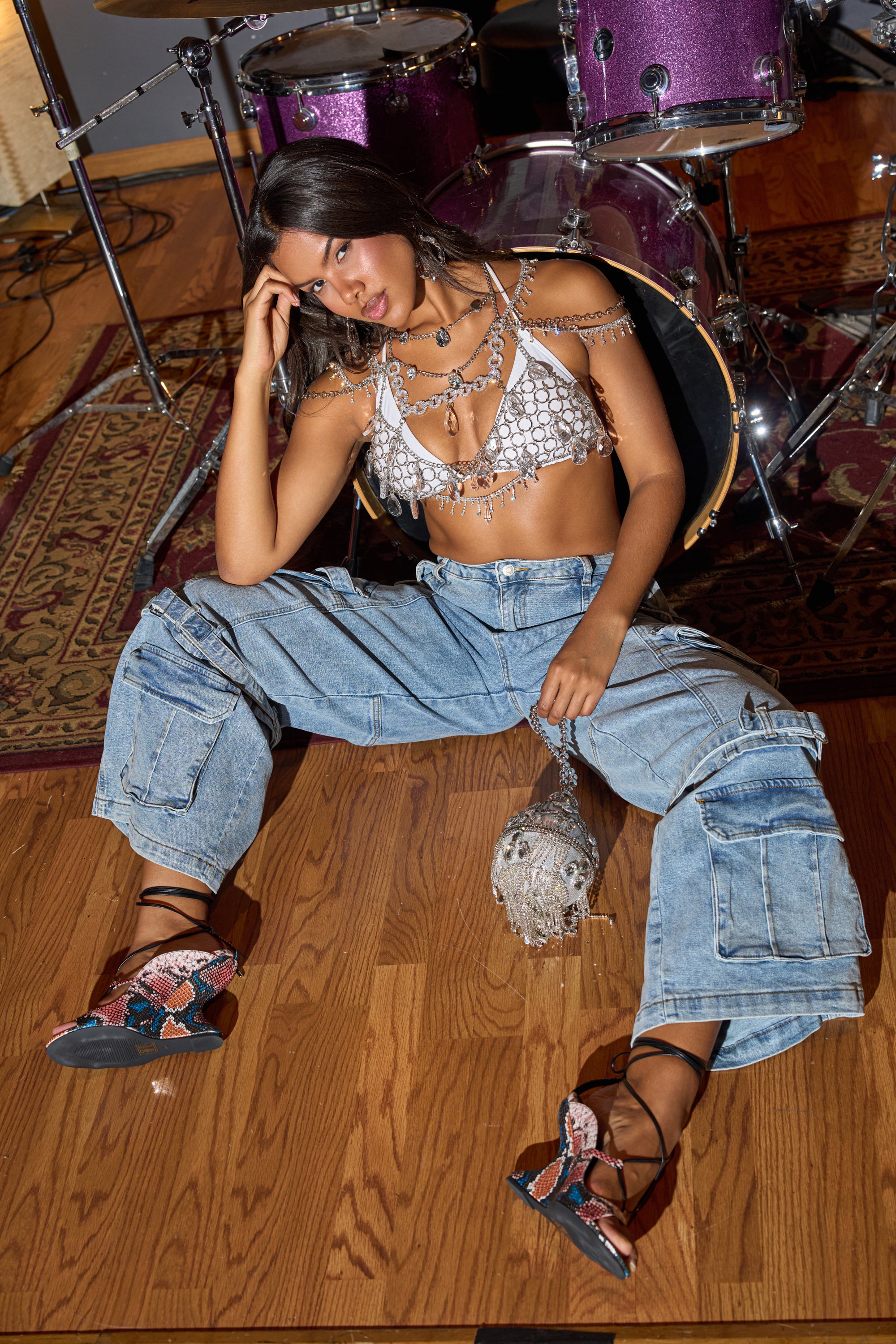A woman sits on a wooden floor with a drum set behind her, wearing the KARA DENIM CARGO PANT, a shiny bralette, layered chains, and open-toe lace-up heels over a patterned rug. 1
