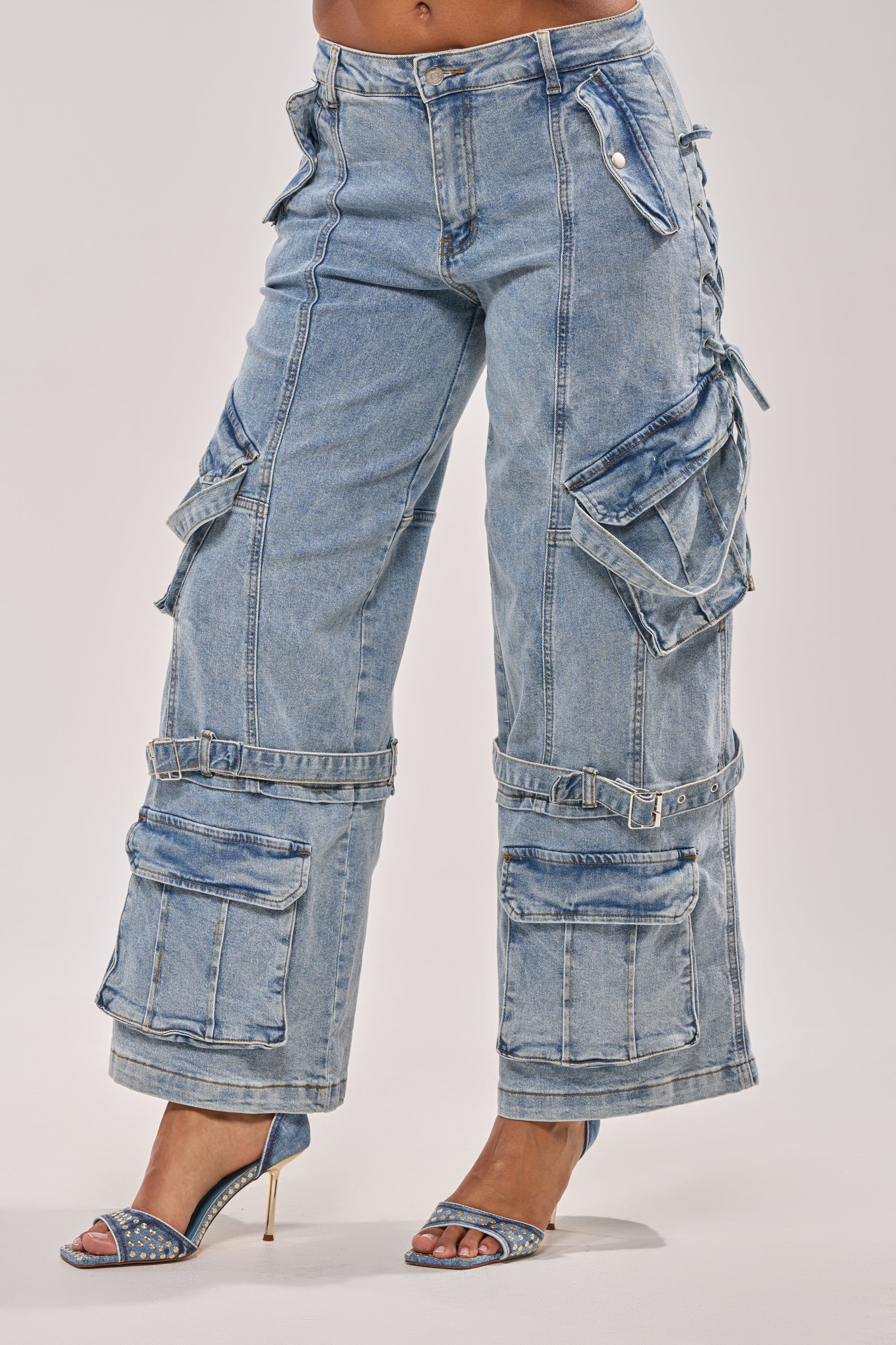 Wearing the KARA DENIM CARGO PANT—light wash denim with adjustable buckles and large pockets—paired with blue rhinestone-studded high-heeled sandals, a person stands against a plain background. 4