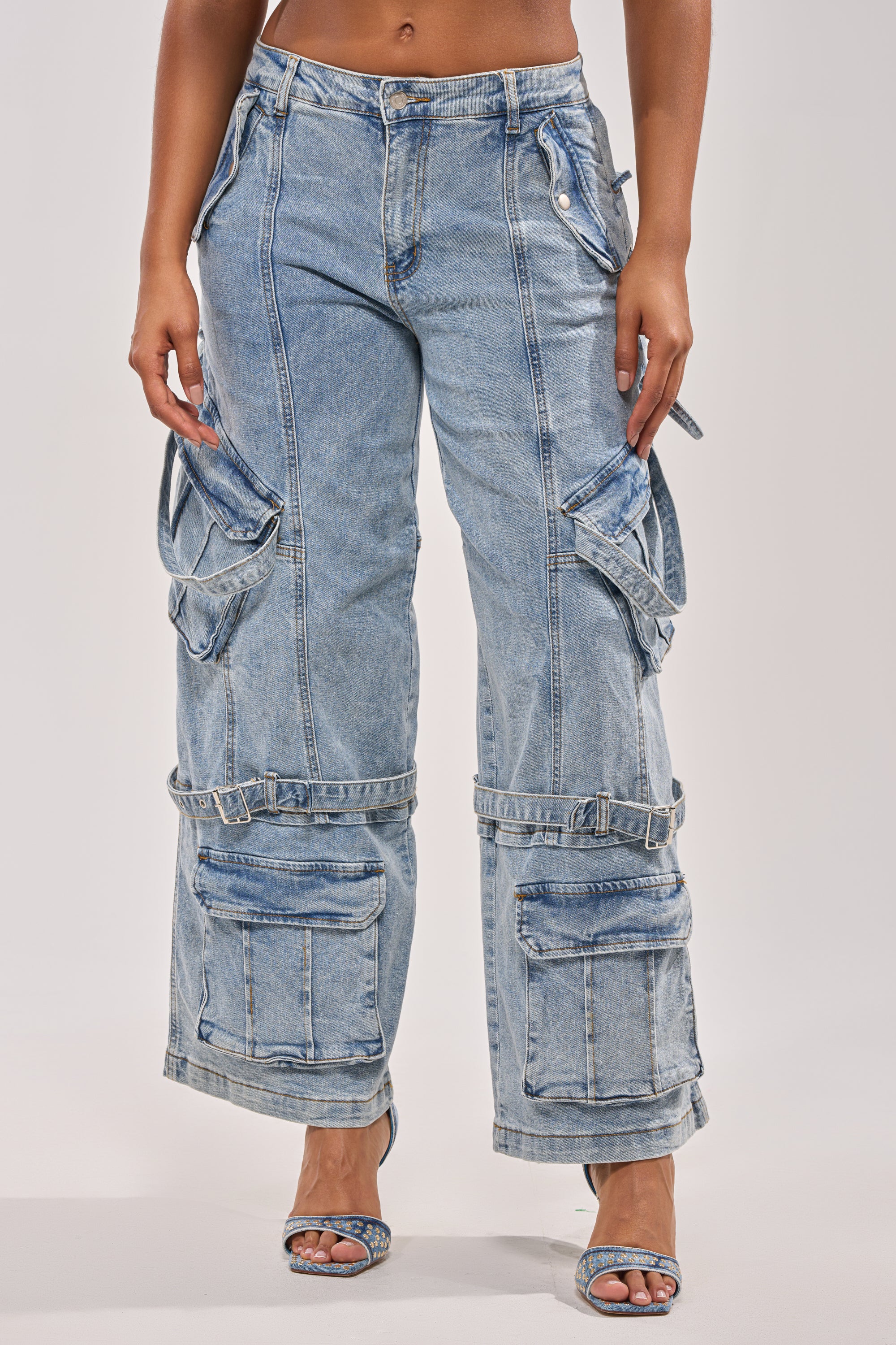 A person wearing KARA DENIM CARGO PANT—a light wash denim cargo jean with adjustable buckle details, multiple large pockets, and straps—paired with open-toe heeled sandals. Image is cropped from the waist down. 5