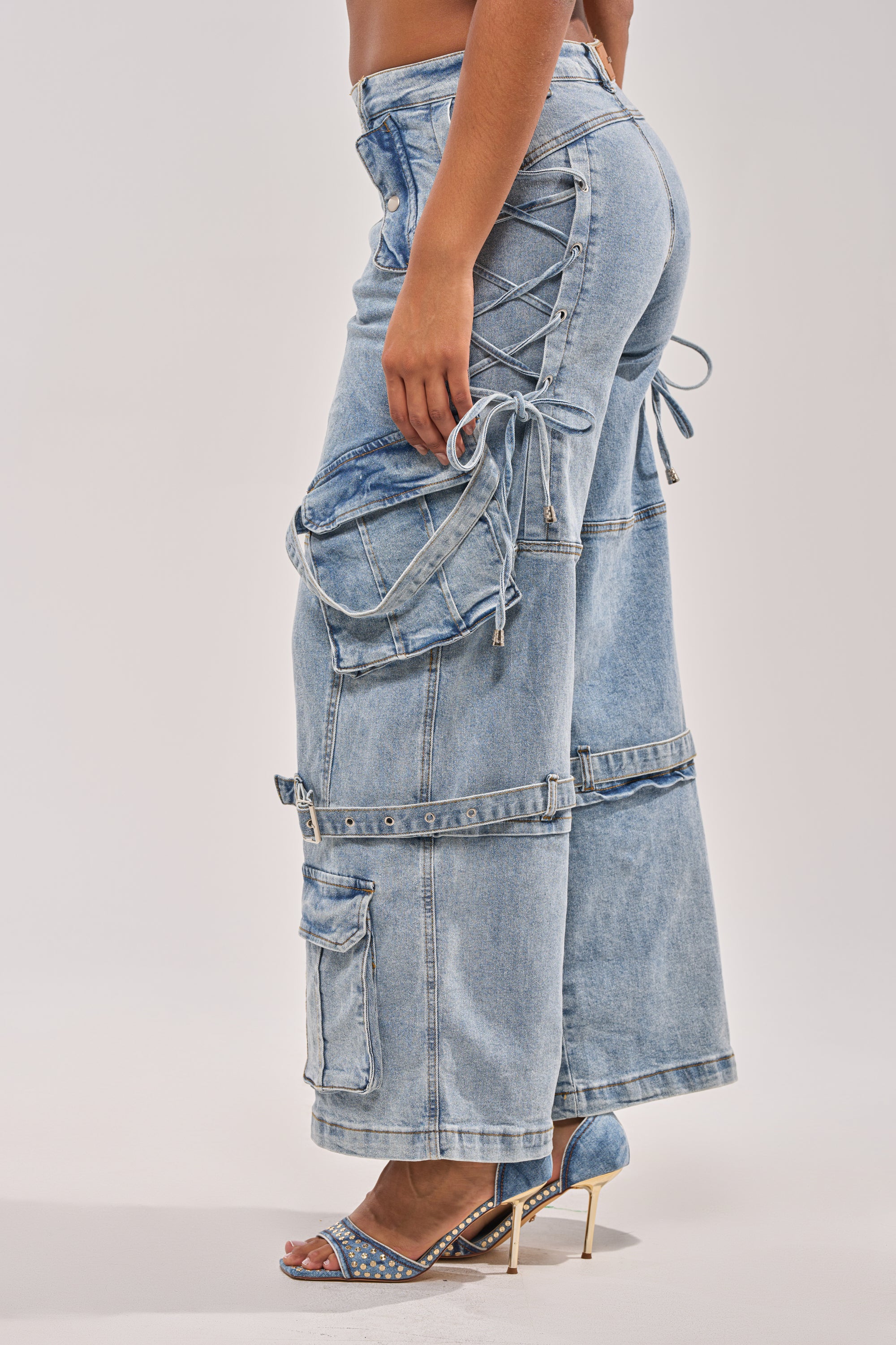 A woman wears the KARA DENIM CARGO PANT, featuring a light wash, lace-up sides, large pockets, and straps, paired with blue high-heeled sandals adorned with rhinestone studs. 7