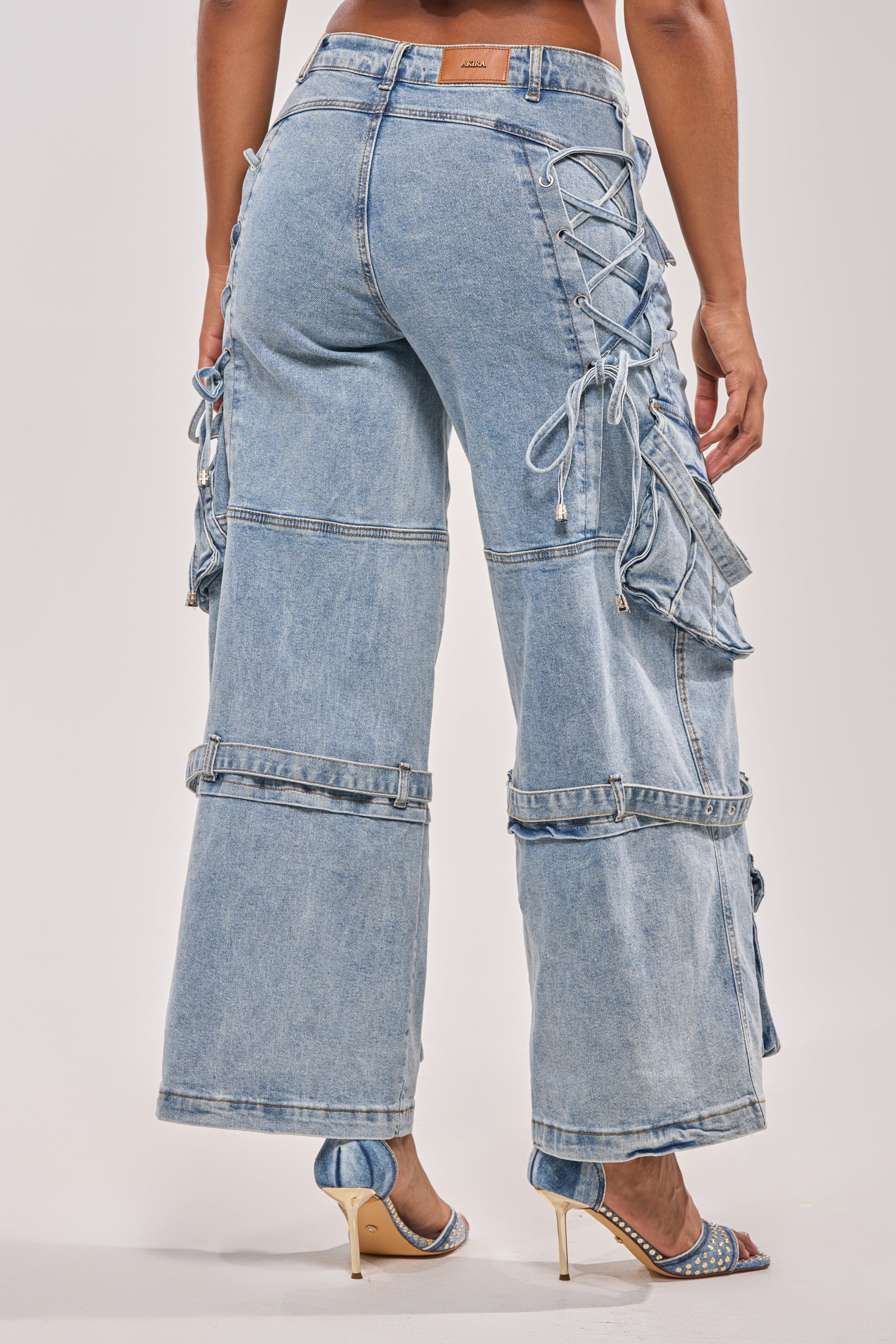 A person is seen from behind wearing KARA DENIM CARGO PANT in light blue with lace-up side details, ruffle accents, and open-toe high heels, highlighting the pants' unique and modern design. 8