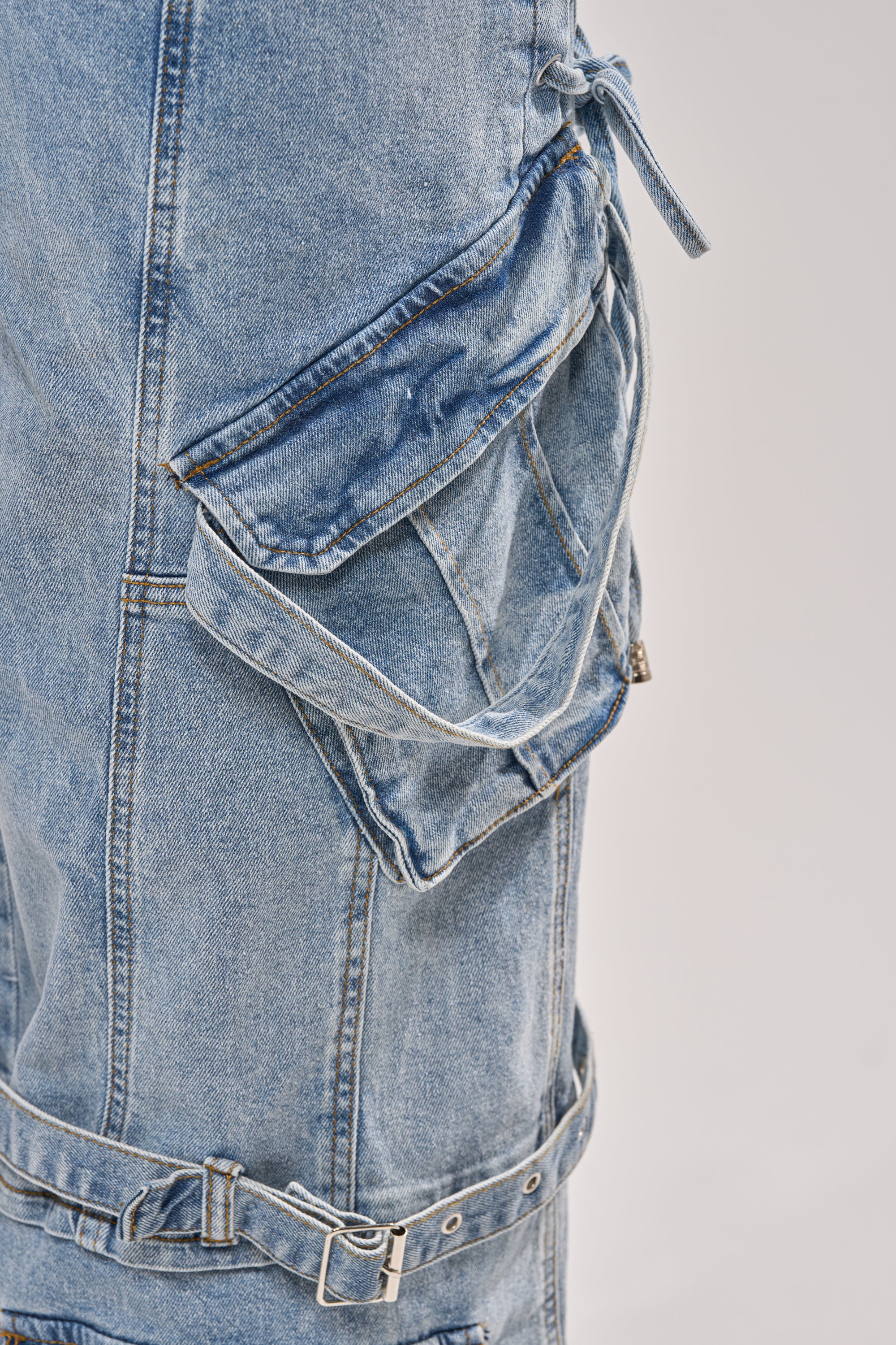 Close-up of the KARA DENIM CARGO PANT in light wash, featuring a large flap pocket with strap detail, visible stitching, and an adjustable buckle denim belt around the thigh. The background is plain and light-colored. 10