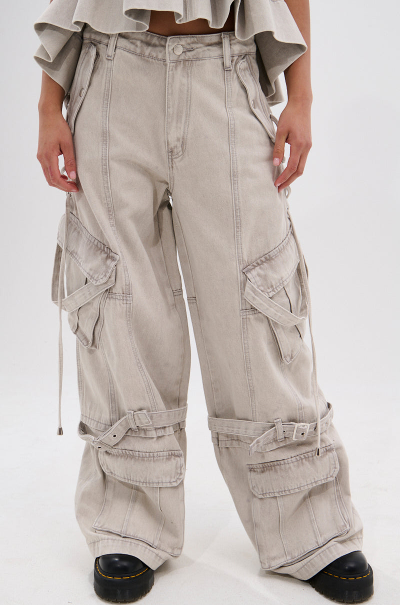 A person wears the KARA DENIM CARGO PANT, featuring a high-waisted, ultra-wide leg silhouette in light gray with multiple pockets, straps, and buckles, paired with black shoes. The image highlights the pants from the waist down.
