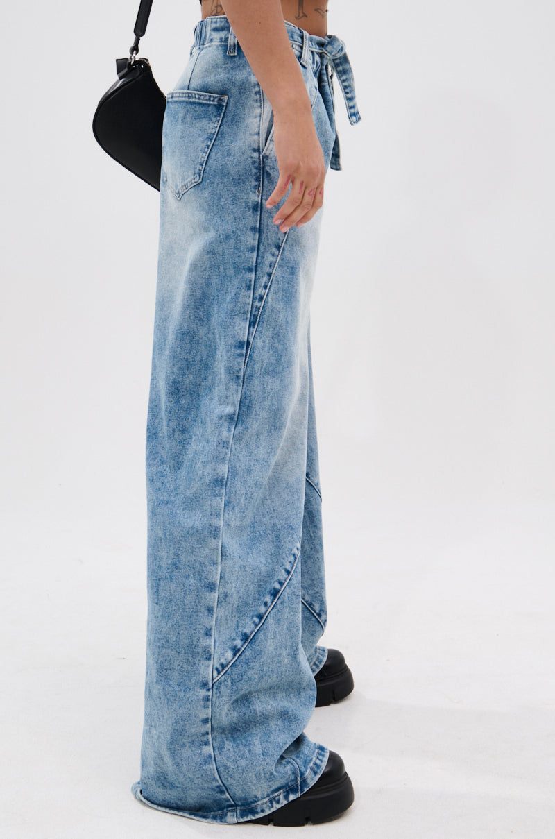Person wears the TIED TO YOU RELAXED DENIM PANT WITH TIE BELT in light wash, styled with black shoes and a black bag. Side view captures the high-rise wide-leg silhouette, loose fit, and unique seam details.