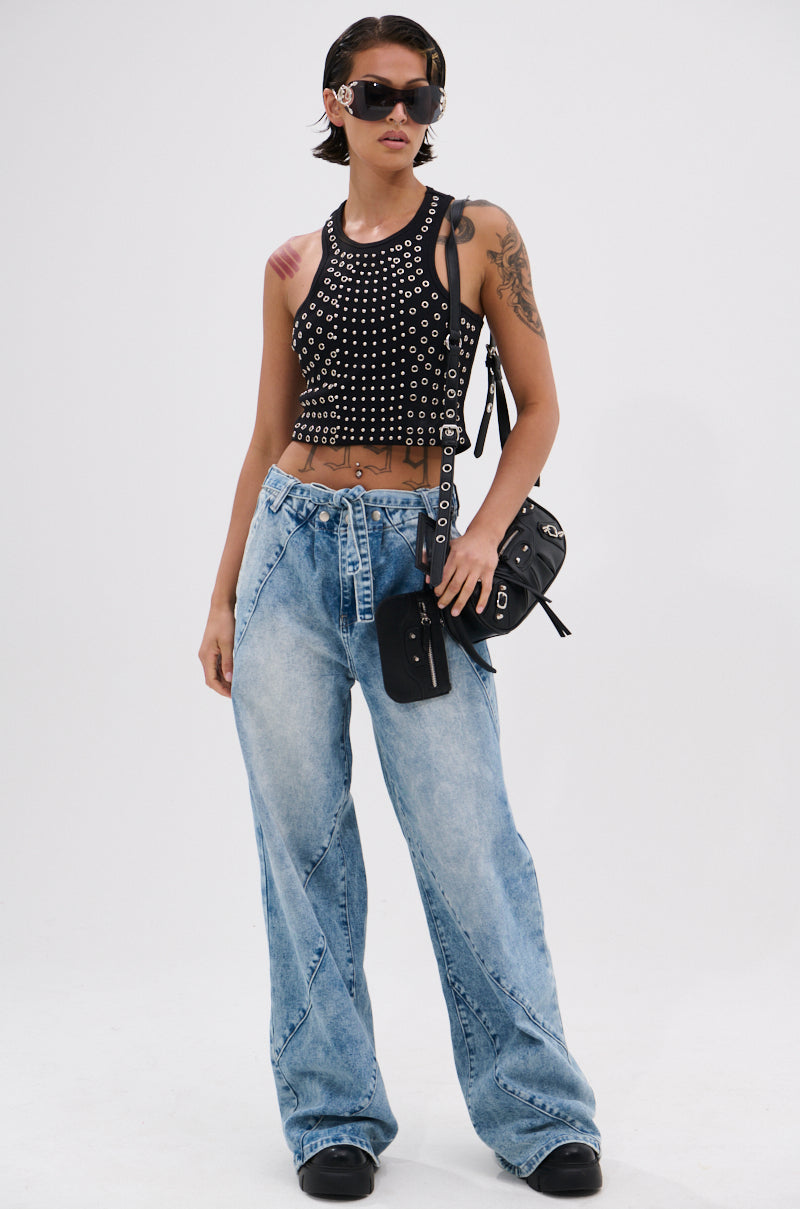 Wearing sunglasses, a black studded crop top, TIED TO YOU RELAXED DENIM PANT WITH TIE BELT, and black platform shoes, a person stands confidently against a plain white background with a small black crossbody bag.