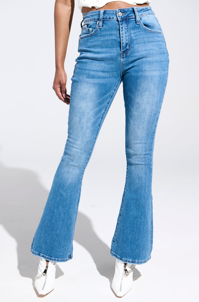 A person stands against a white background wearing RIP YOU OFF BOOTCUT JEANS, high-waisted and light blue with a slightly faded look, complemented by white ankle boots. The semi-stretchy denim ensures a comfortable fit, enhancing the jeans' timeless appeal.