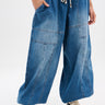 Model wears the DONT MESS WITH ME DENIM JOGGER—high-rise blue denim cargo joggers with large front pockets and a white drawstring—styled with chunky black streetwear shoes with beige laces, standing against a plain white background.