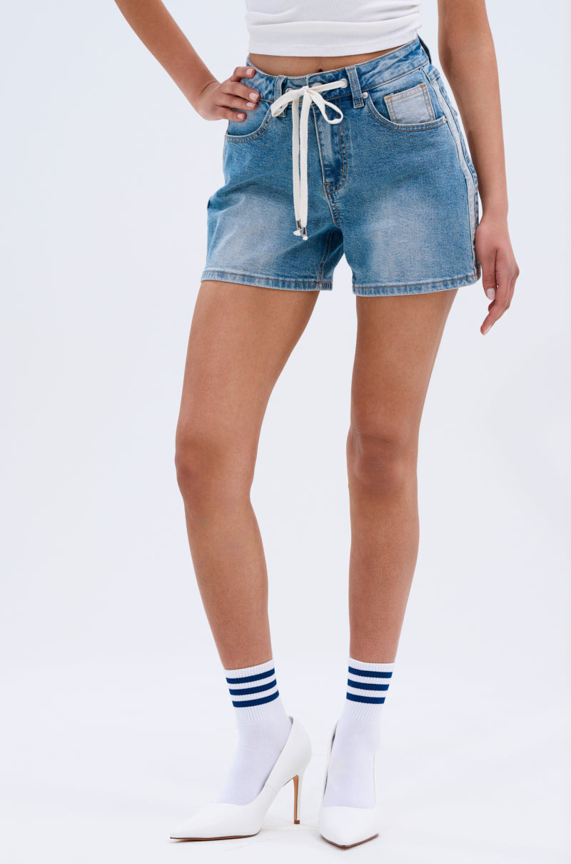 Wearing the REMIND ME DENIM SHORT in vintage blue with a white drawstring, paired with a white sleeveless top, white socks with blue stripes, and white high-heeled shoes, standing against a plain light background.