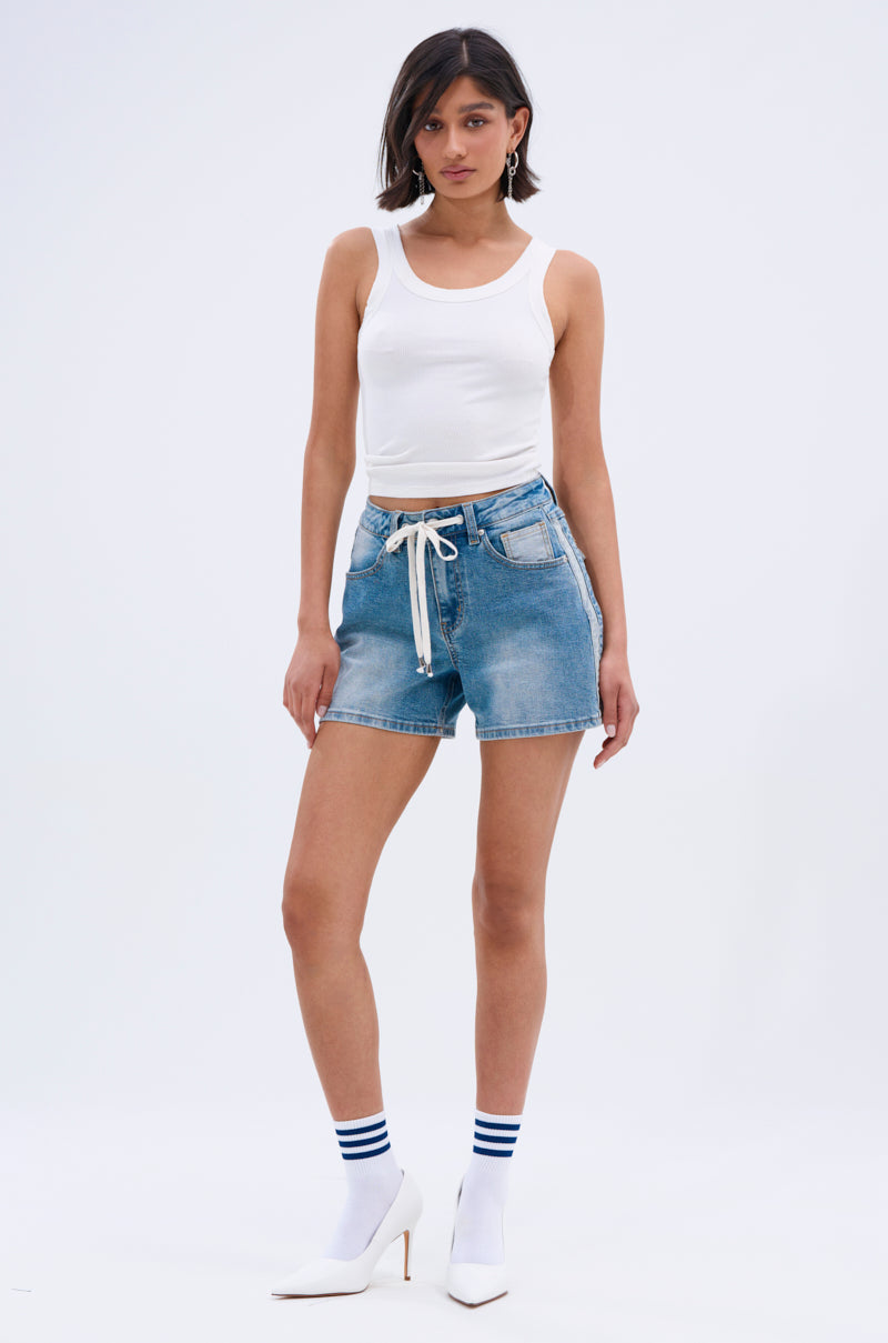 A woman with short dark hair and gold hoop earrings wears a white tank top, REMIND ME DENIM SHORT in vintage blue wash with a white drawstring, white heeled ankle boots, and white socks with blue stripes against a plain background.