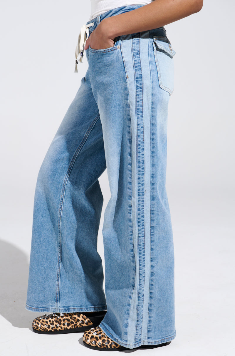 A person wears the JUST LIKE THAT WIDE LEG DENIM PANT with a vintage fade, side stripe, white top, and leopard print shoes, standing sideways against a plain white background.