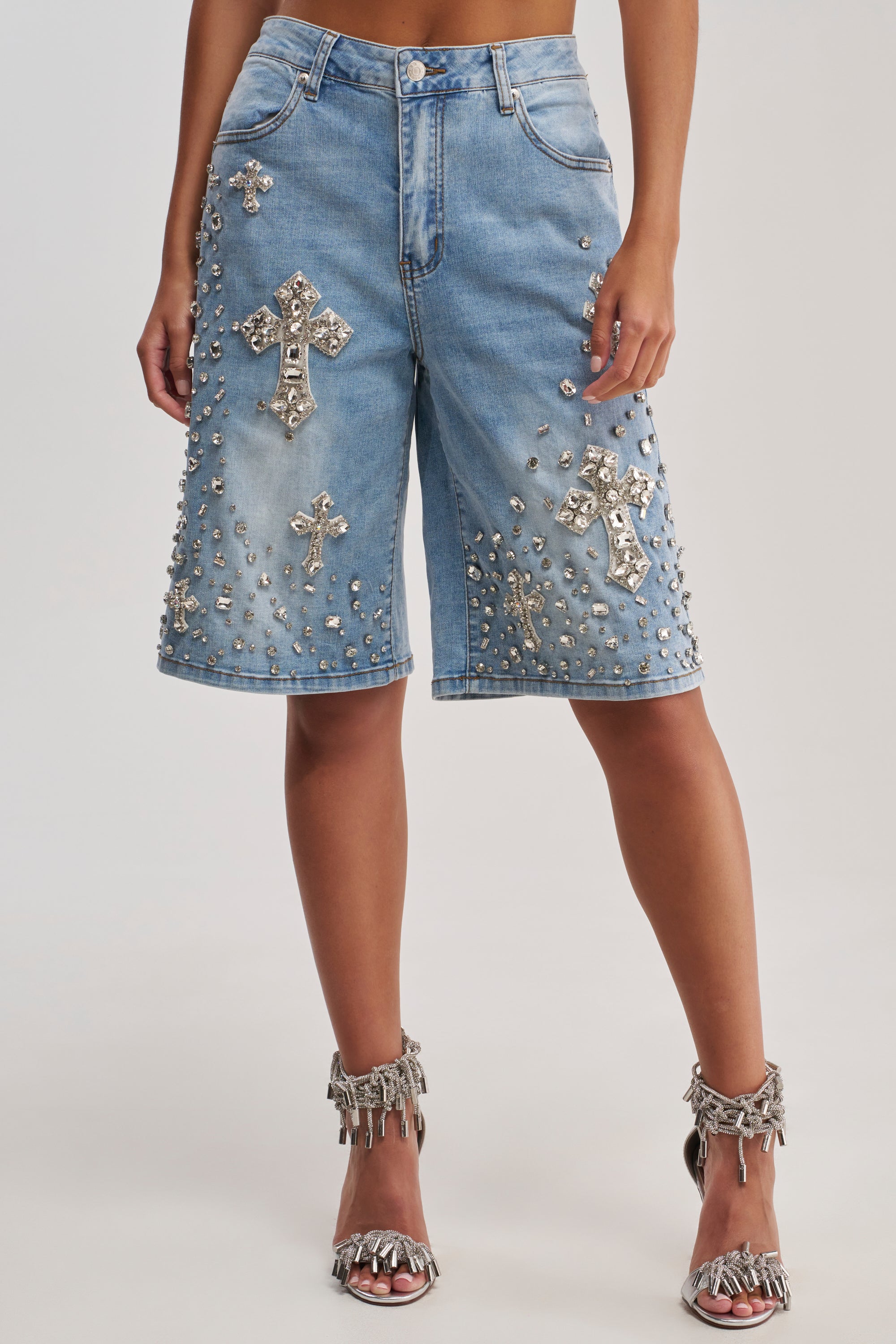 A person wearing the BLESSED RHINESTONE BERMUDA, featuring knee-length denim embellished with large silver jeweled crosses and studs, styled with silver fringed high-heeled sandals.